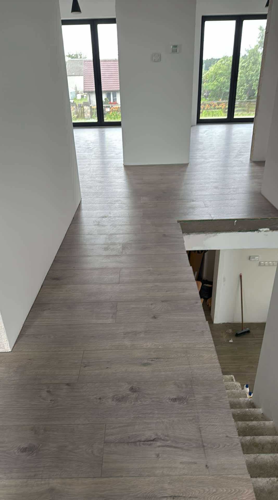 Ułożony parkiet w jasnym odcieniu drewna w nowoczesnym wnętrzu domu z dużymi oknami i schodami. Widoczny fragment pomieszczenia poniżej schodów.