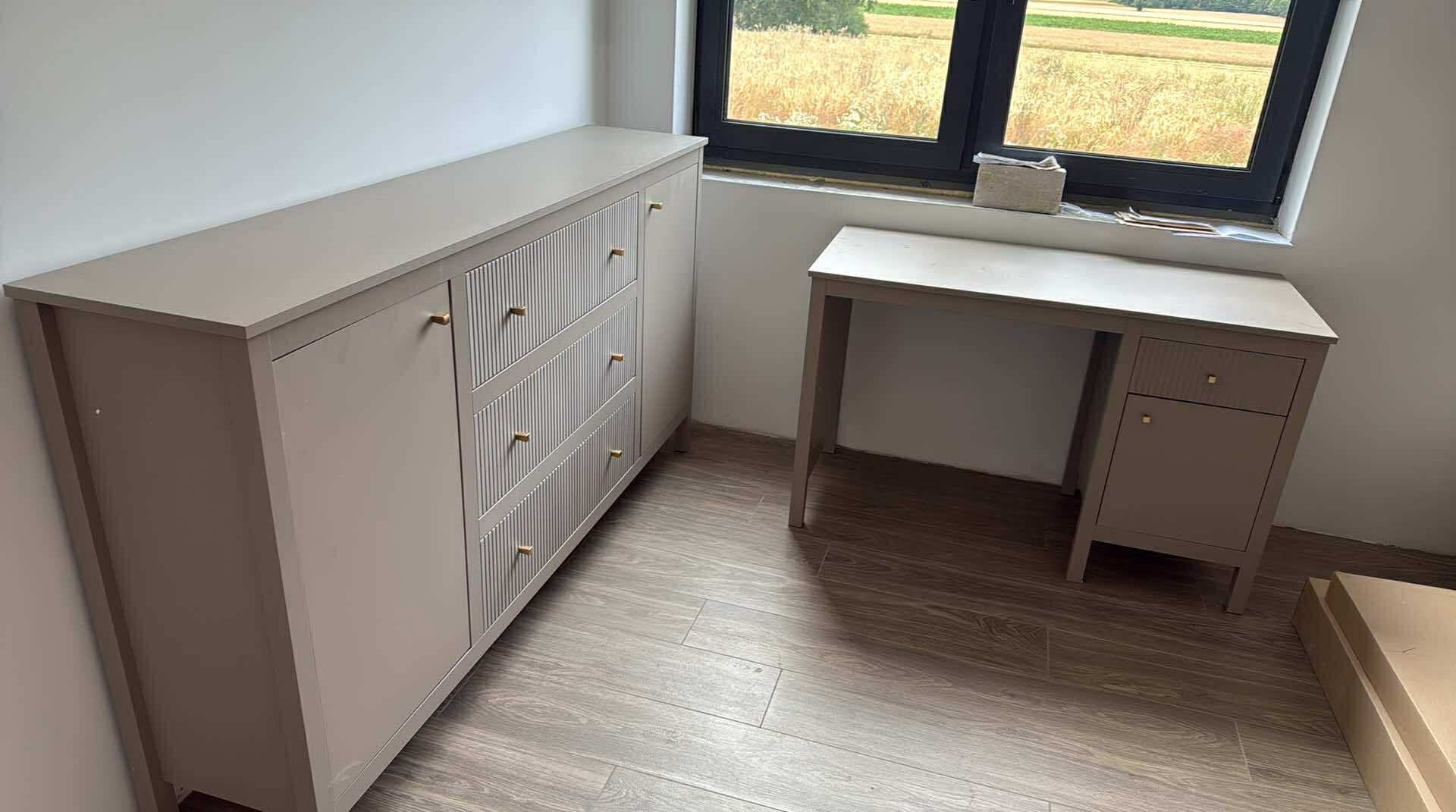 Beżowy zestaw mebli z ryflowanymi frontami szuflad i złotymi uchwytami, stojący na panelach podłogowych w pokoju z oknem wychodzącym na pola.