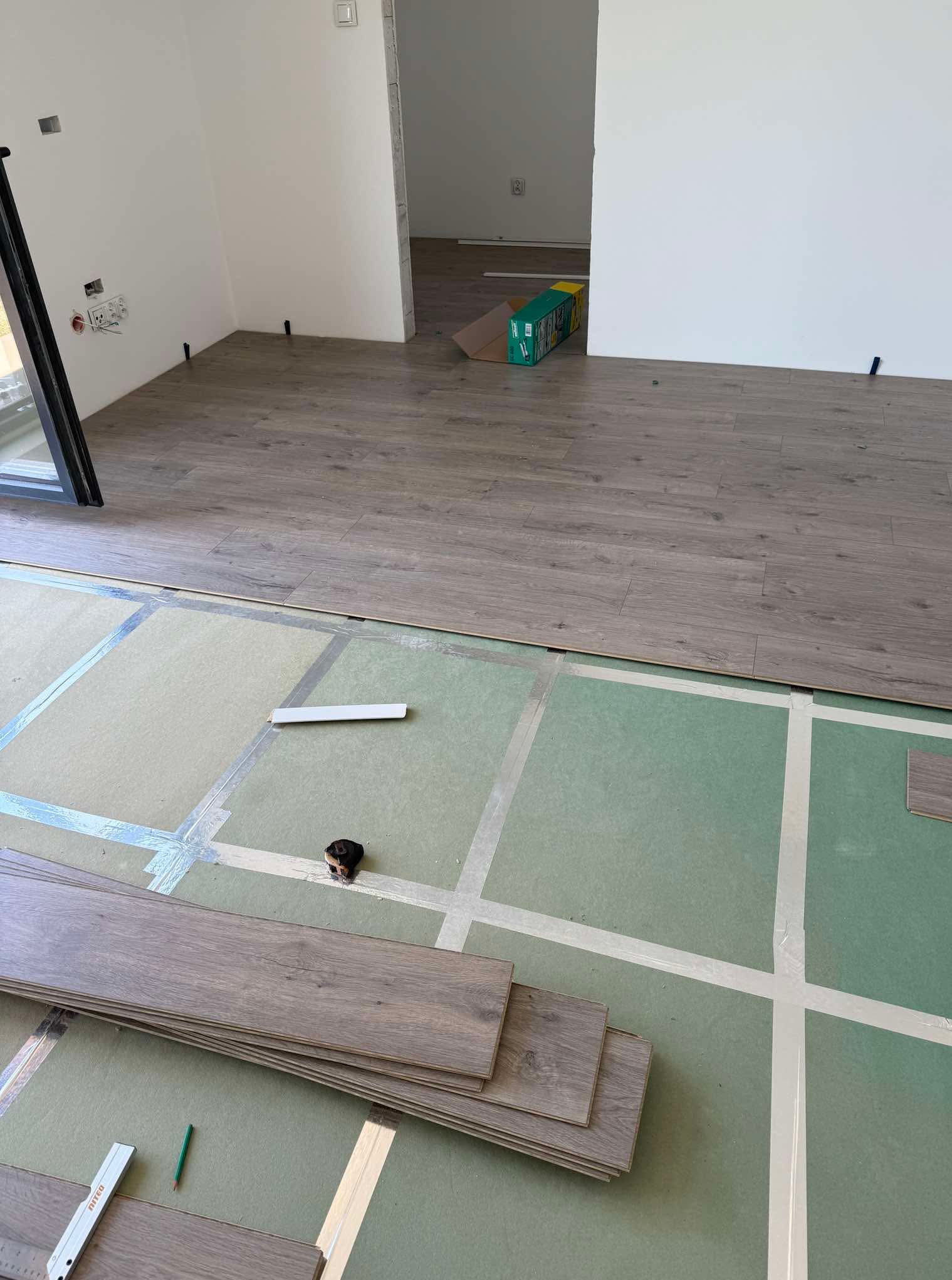 Układanie paneli podłogowych: widoczne panele, podkład, taśma miernicza i ołówek. Częściowo wykończona podłoga w jasnym pomieszczeniu.