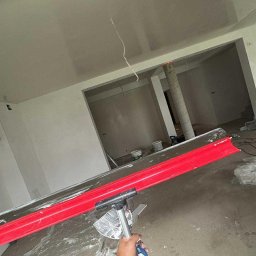 SŁOWIK INTERIORS - Maciej Słowik - Świeżo wygładzona powierzchnia sufitu w jasnym wnętrzu, widoczna z perspektywy osoby trzymającej narzędzie do rozprowadzania masy. W tle surowe ściany i kolumna.