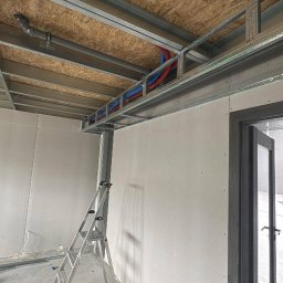 SŁOWIK INTERIORS - Maciej Słowik - Montaż sufitu podwieszanego z płyt OSB i instalacji. Widoczne metalowe profile, rury instalacyjne i drabina. Ściany przygotowane do dalszych prac.