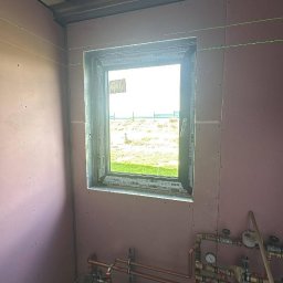 SŁOWIK INTERIORS - Maciej Słowik - Wnętrze w trakcie prac: ściany z płyt gipsowo-kartonowych, widoczne instalacje grzewcze, okno z widokiem na zewnątrz, metalowa konstrukcja sufitu i poziomica laserowa.