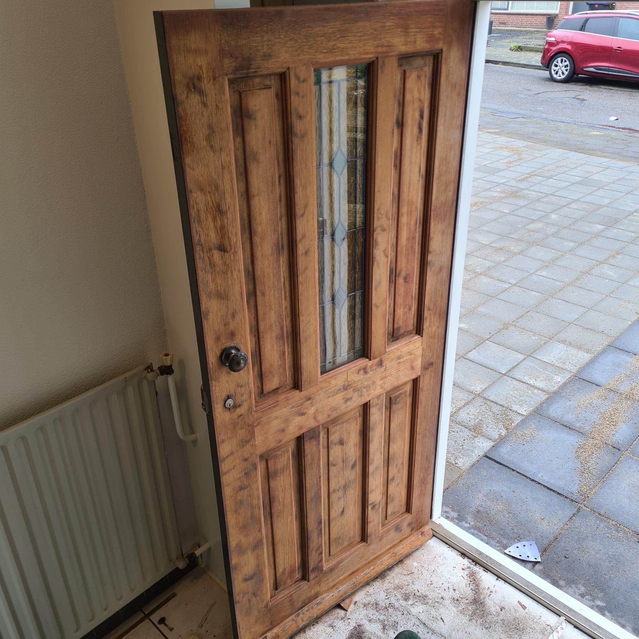 Drewniane drzwi wejściowe z szybą po renowacji, widoczne ślady szlifowania, na zewnątrz kostka brukowa i czerwony samochód. Detal z bliska, ciepłe oświetlenie.