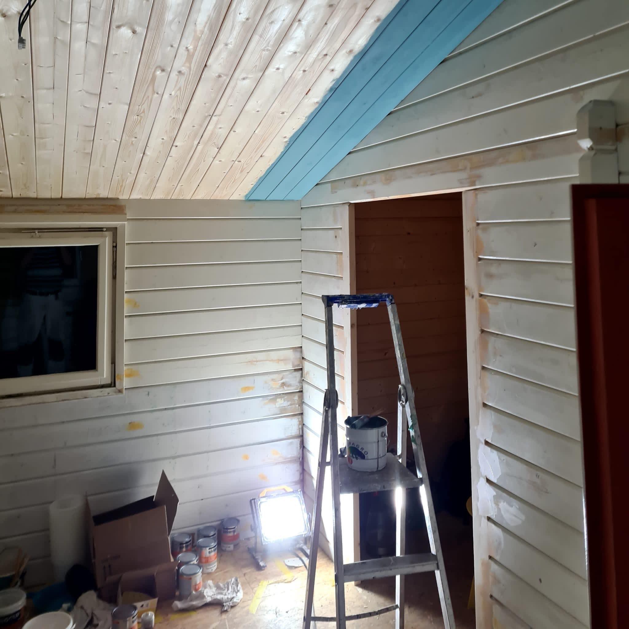 Wnętrze w trakcie malowania: drewniana boazeria częściowo pomalowana na biało, fragment sufitu na niebiesko. Widoczna drabina, puszki z farbą i lampa robocza.