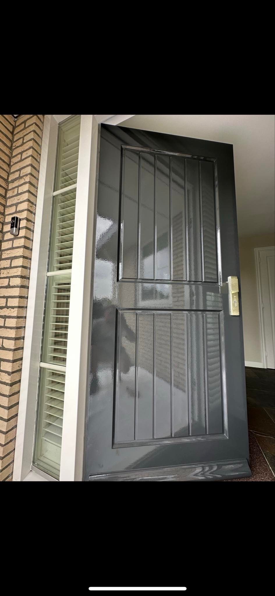 Szare, lakierowane drzwi wejściowe z metalowymi wstawkami, zamontowane obok okna z żaluzjami, widoczne z bliska pod kątem.