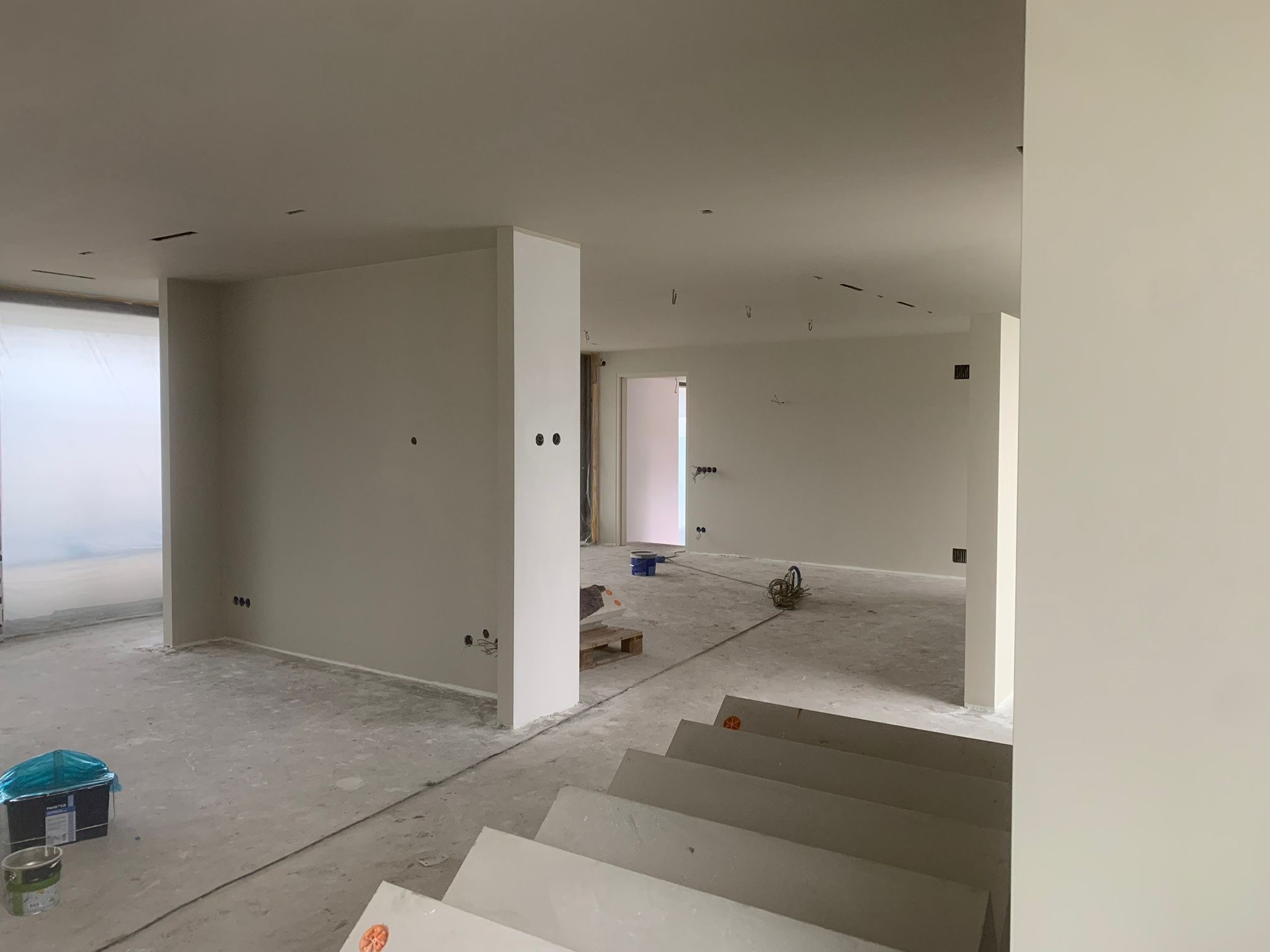 Wnętrze w trakcie prac wykończeniowych: świeżo pomalowane ściany w jasnym odcieniu, widoczne puszki elektryczne, podłoga przygotowana do dalszych prac. Schody w surowym stanie.