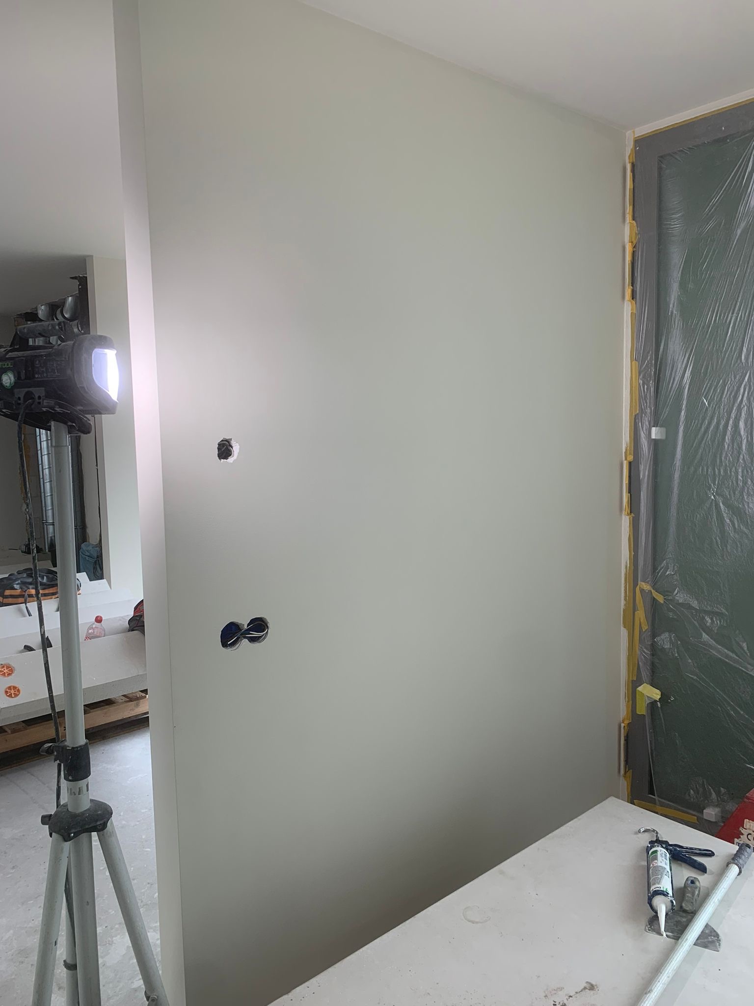 Świeżo pomalowana ściana w jasnym odcieniu z widocznymi otworami na gniazdka elektryczne, oświetlona lampą budowlaną. Okno zabezpieczone folią malarską.