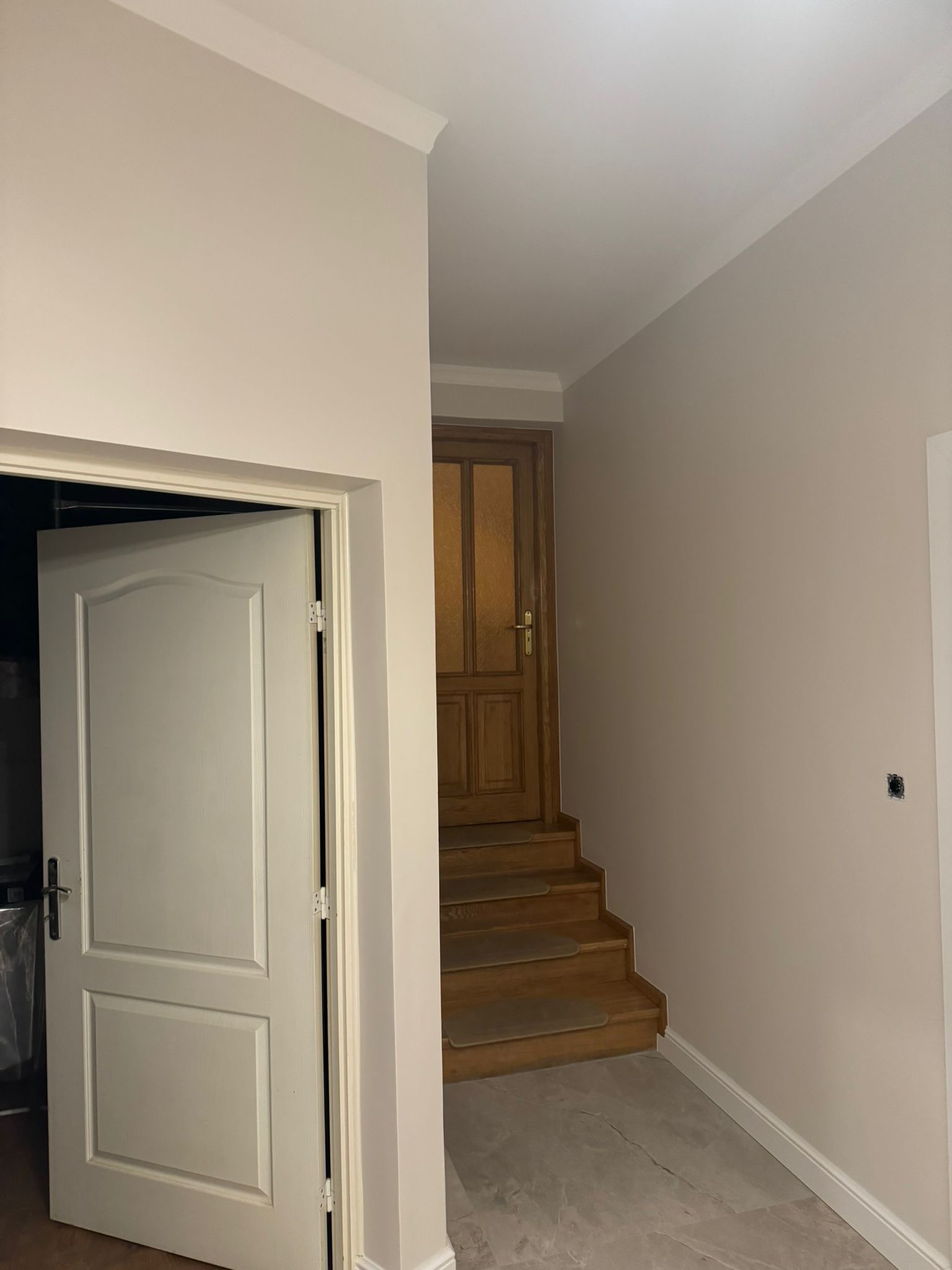 Jasne wnętrze po remoncie: pomalowane ściany w odcieniach beżu, białe listwy przypodłogowe, drewniane schody i dwoje drzwi, jedne otwarte, drugie prowadzące na schody.