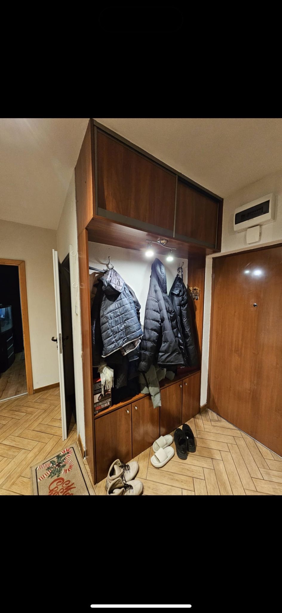 Drewniana zabudowa przedpokoju z szafkami, wieszakami i oświetleniem punktowym. Na podłodze leżą buty i kapcie, a obok wejścia dywanik.