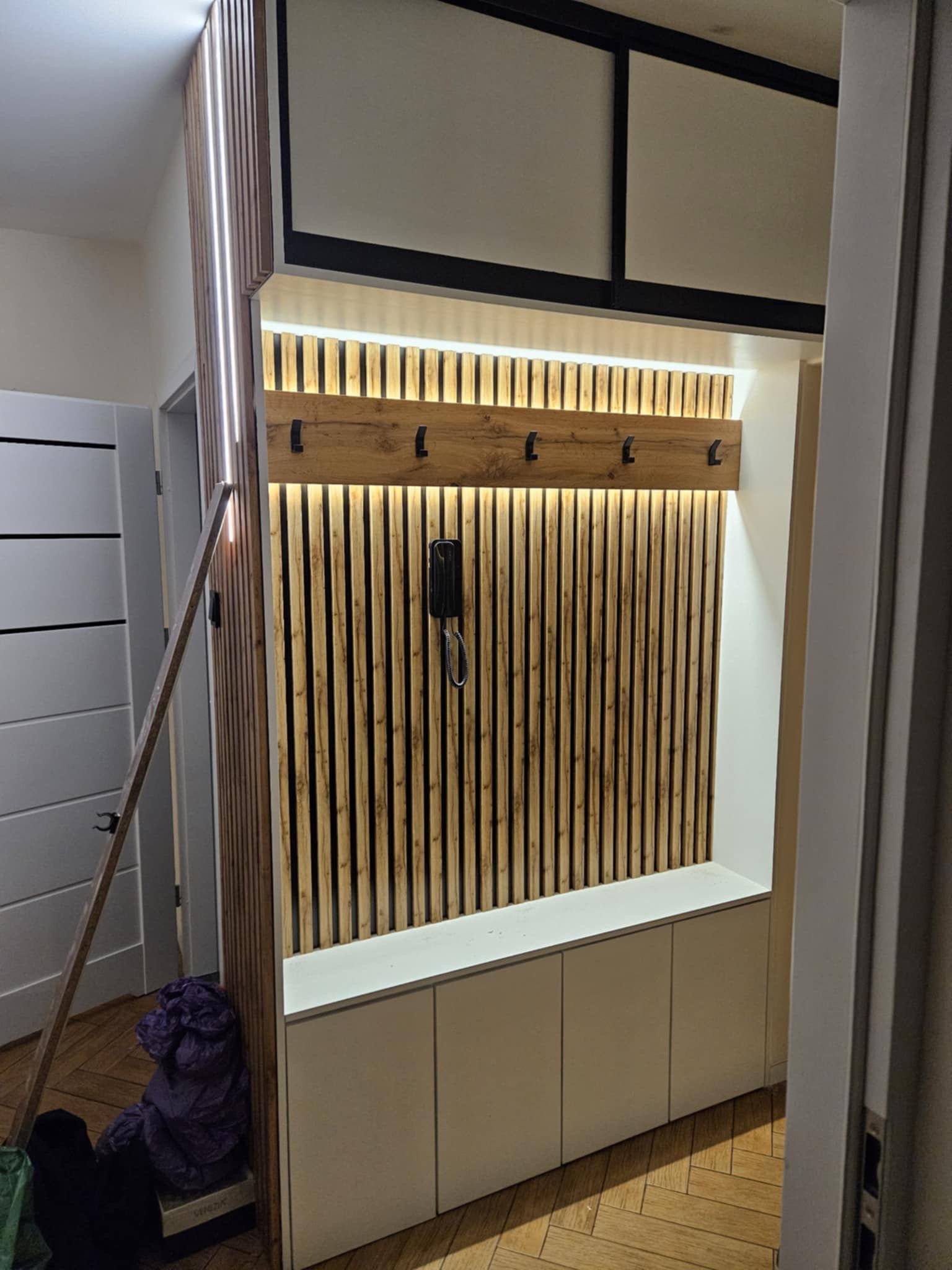 Nowoczesna zabudowa przedpokoju z drewnianymi lamelami i oświetleniem LED, wnęka z szafkami i wieszakami, parkiet w jodełkę. Wykończenie wnętrza w jasnych barwach.