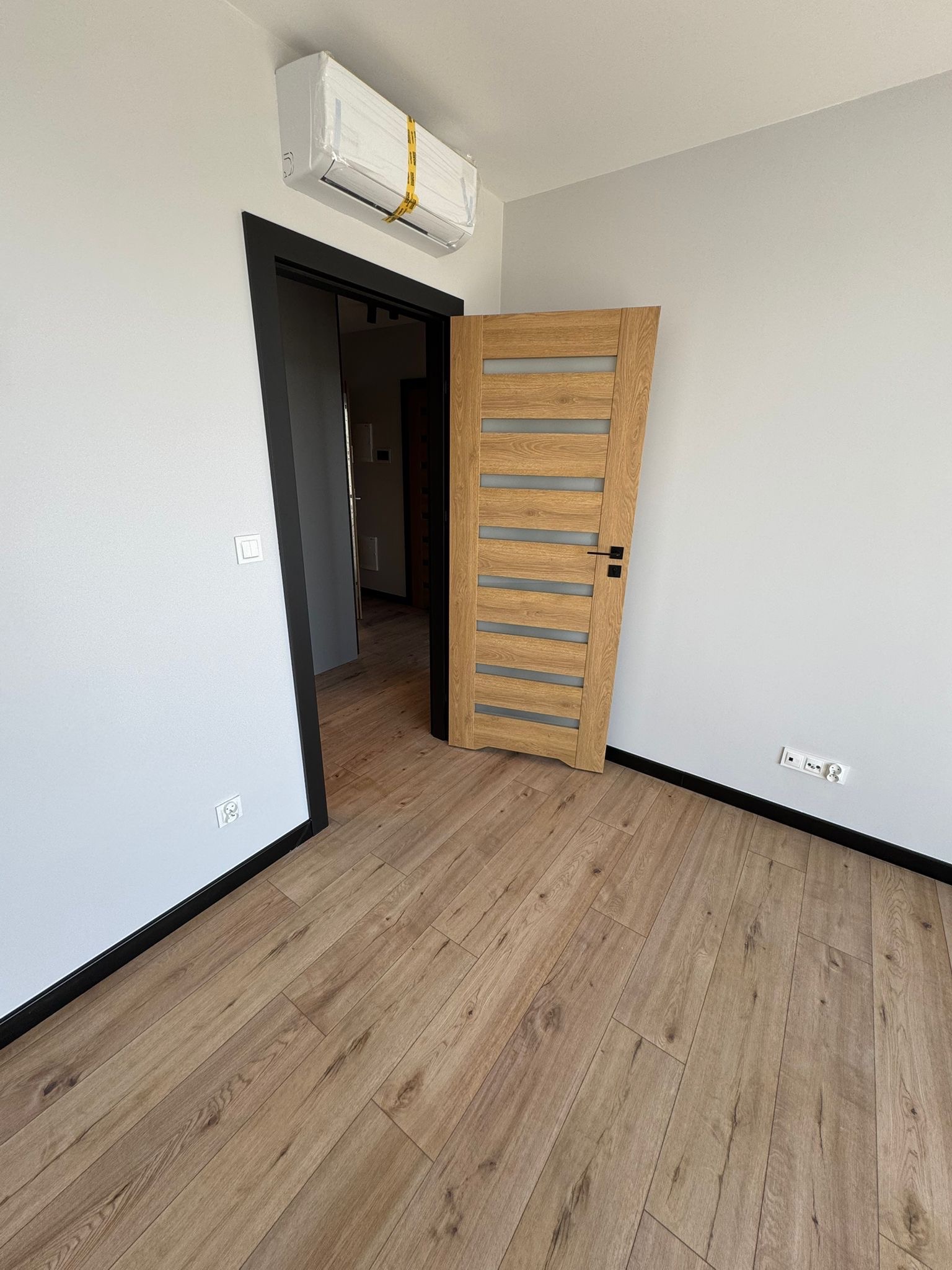 Jasne panele podłogowe w nowym wnętrzu z drewnianymi drzwiami z przeszkleniami i klimatyzatorem w folii. Świeżo pomalowane ściany i czarne listwy przypodłogowe.