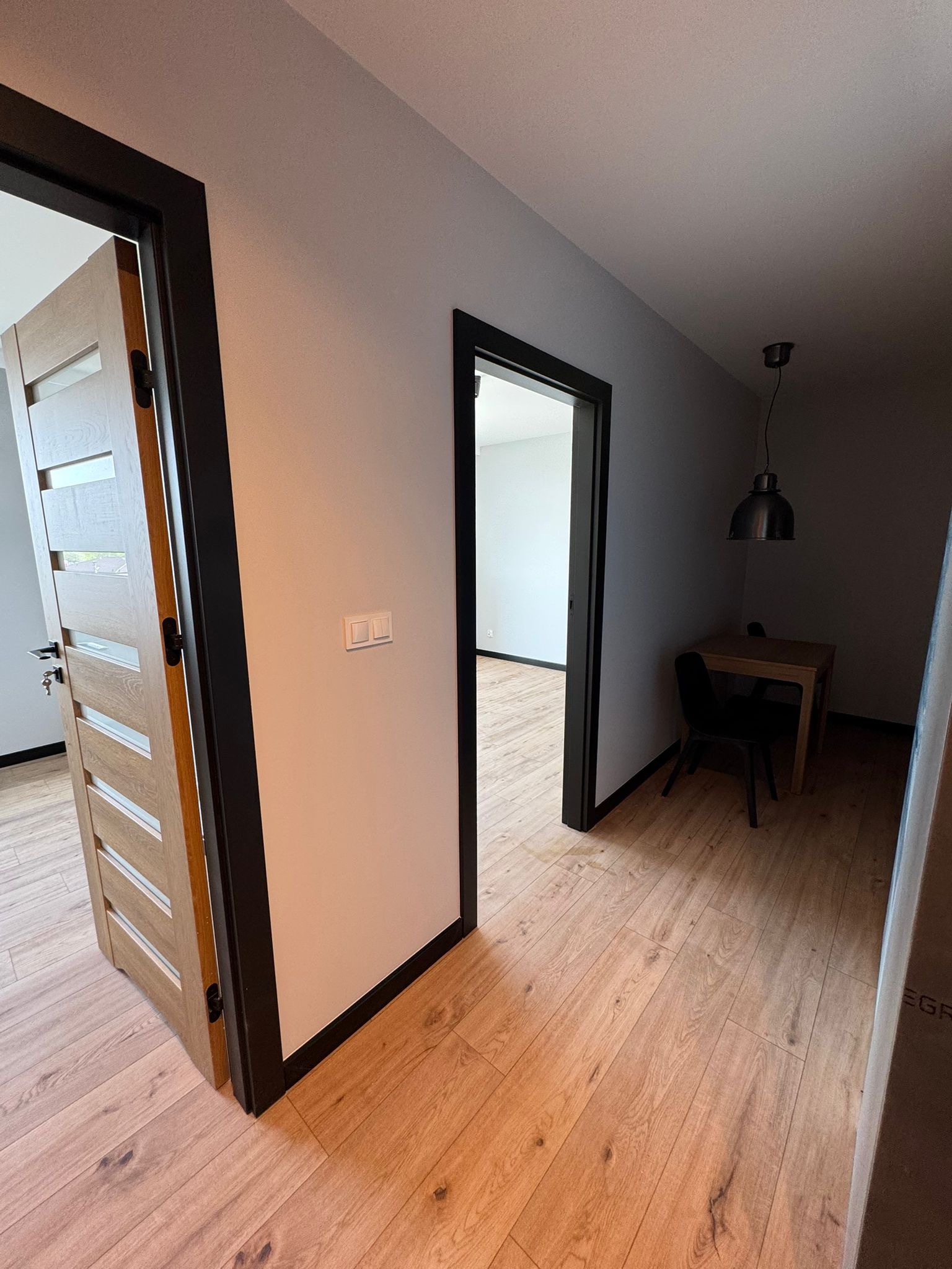 Jasne panele podłogowe, białe ściany z czarnymi obramowaniami drzwi. Otwarte drzwi z drewnianymi panelami i widok na wnętrze z stołem i krzesłem.