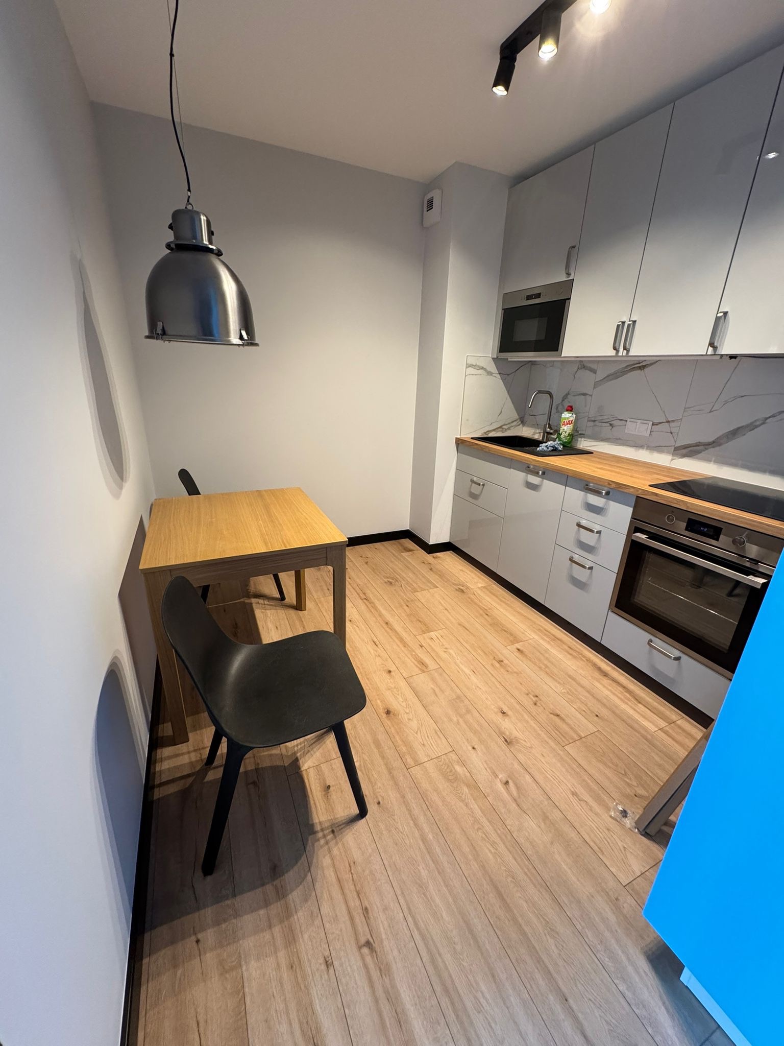 Jasna kuchnia z drewnianym blatem i podłogą, szare szafki, marmurowy backsplash, stół z krzesłem. Nowoczesne oświetlenie sufitowe i lampa wisząca.