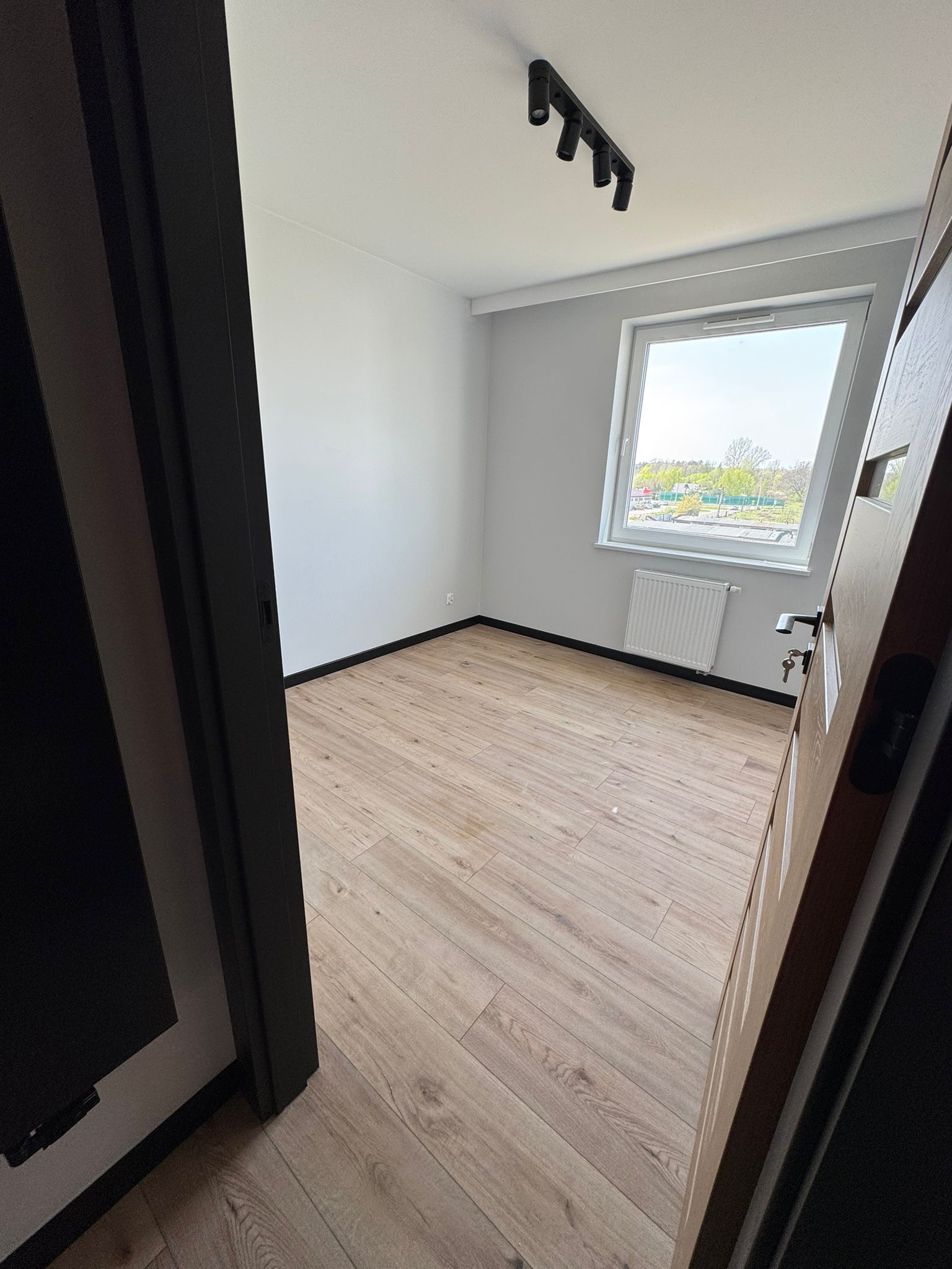 Jasne, wykończone wnętrze pokoju z drewnianą podłogą, białymi ścianami i oknem z widokiem na zieleń. Czarna lampa sufitowa i otwarte drzwi z kluczami.