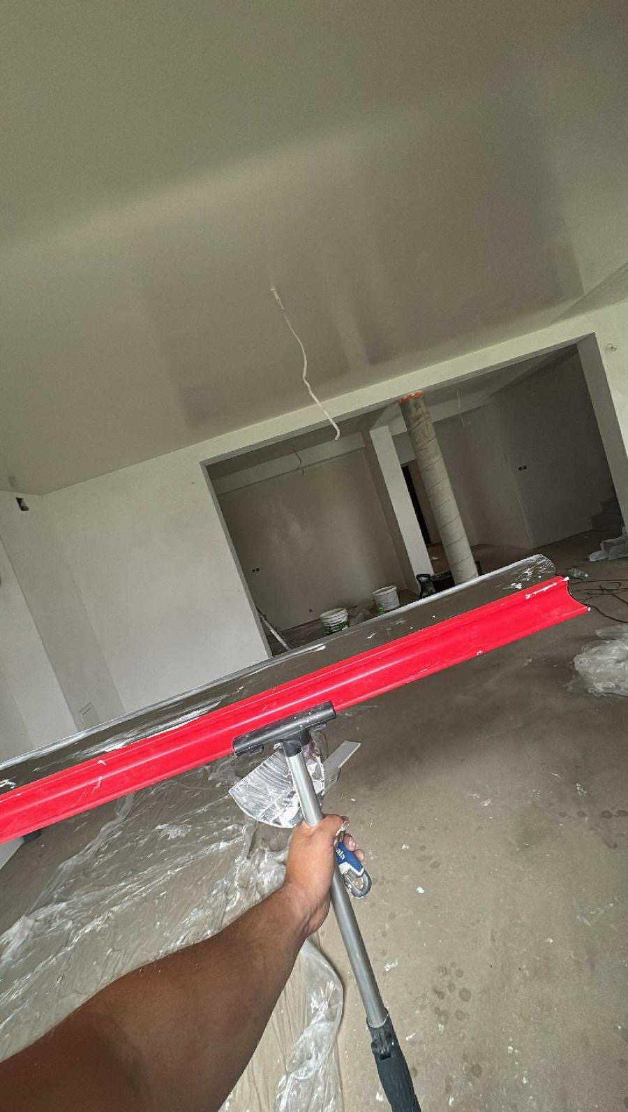 Świeżo wygładzona powierzchnia sufitu w jasnym wnętrzu, widoczna z perspektywy osoby trzymającej narzędzie do rozprowadzania masy. W tle surowe ściany i kolumna.