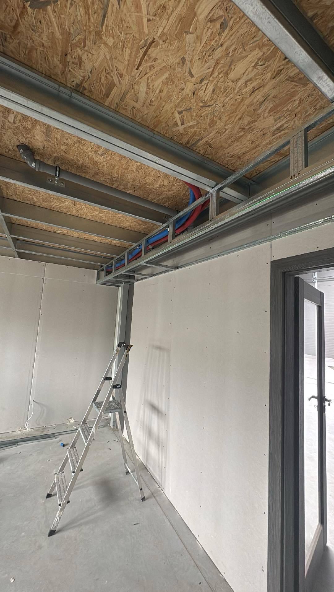 Montaż sufitu podwieszanego z płyt OSB i instalacji. Widoczne metalowe profile, rury instalacyjne i drabina. Ściany przygotowane do dalszych prac.