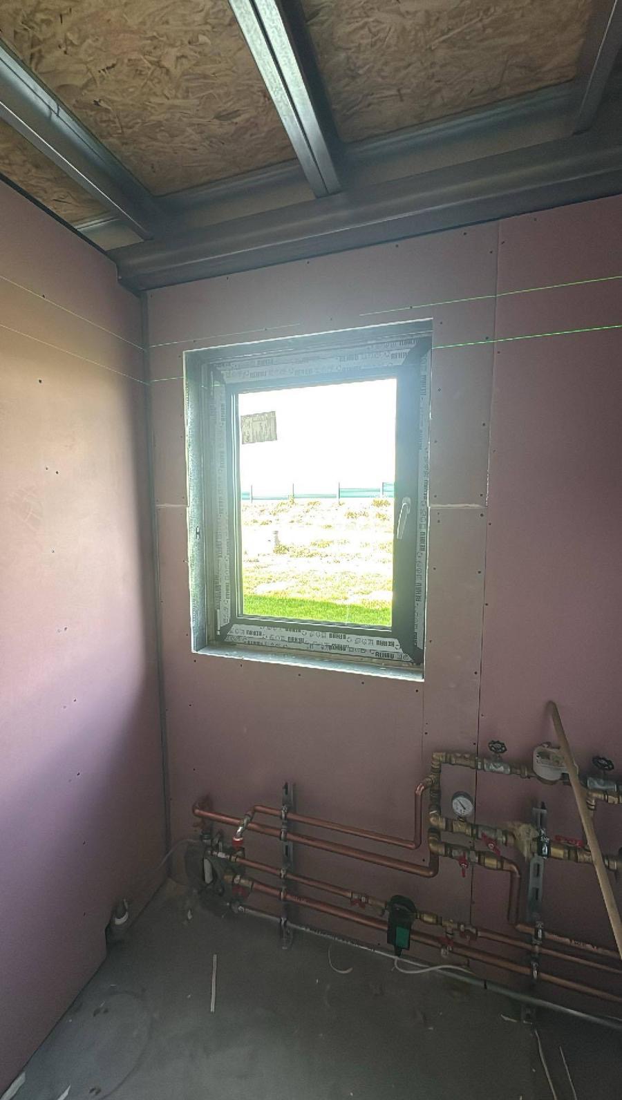 Wnętrze w trakcie prac: ściany z płyt gipsowo-kartonowych, widoczne instalacje grzewcze, okno z widokiem na zewnątrz, metalowa konstrukcja sufitu i poziomica laserowa.