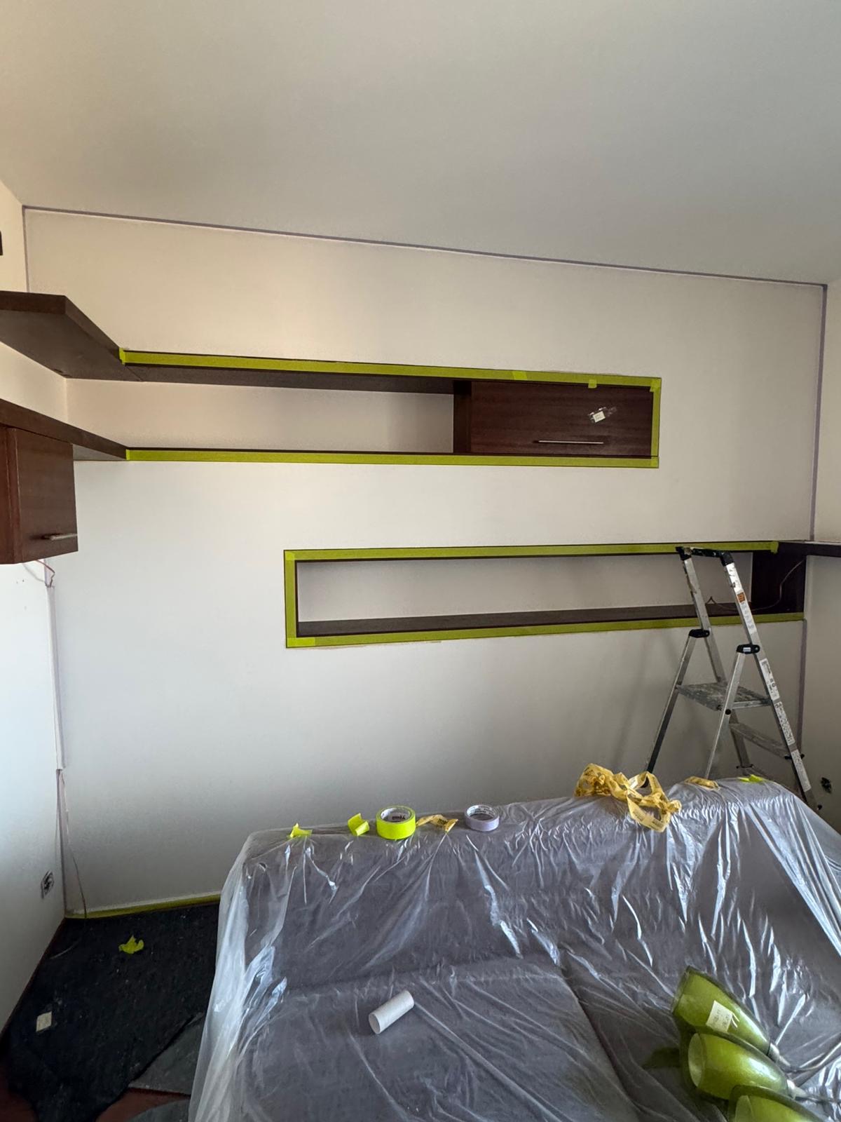 Biała ściana z zamontowanymi półkami i szafkami, zabezpieczona taśmą malarską. Sofa przykryta folią ochronną, drabina w tle. Przygotowania do malowania.