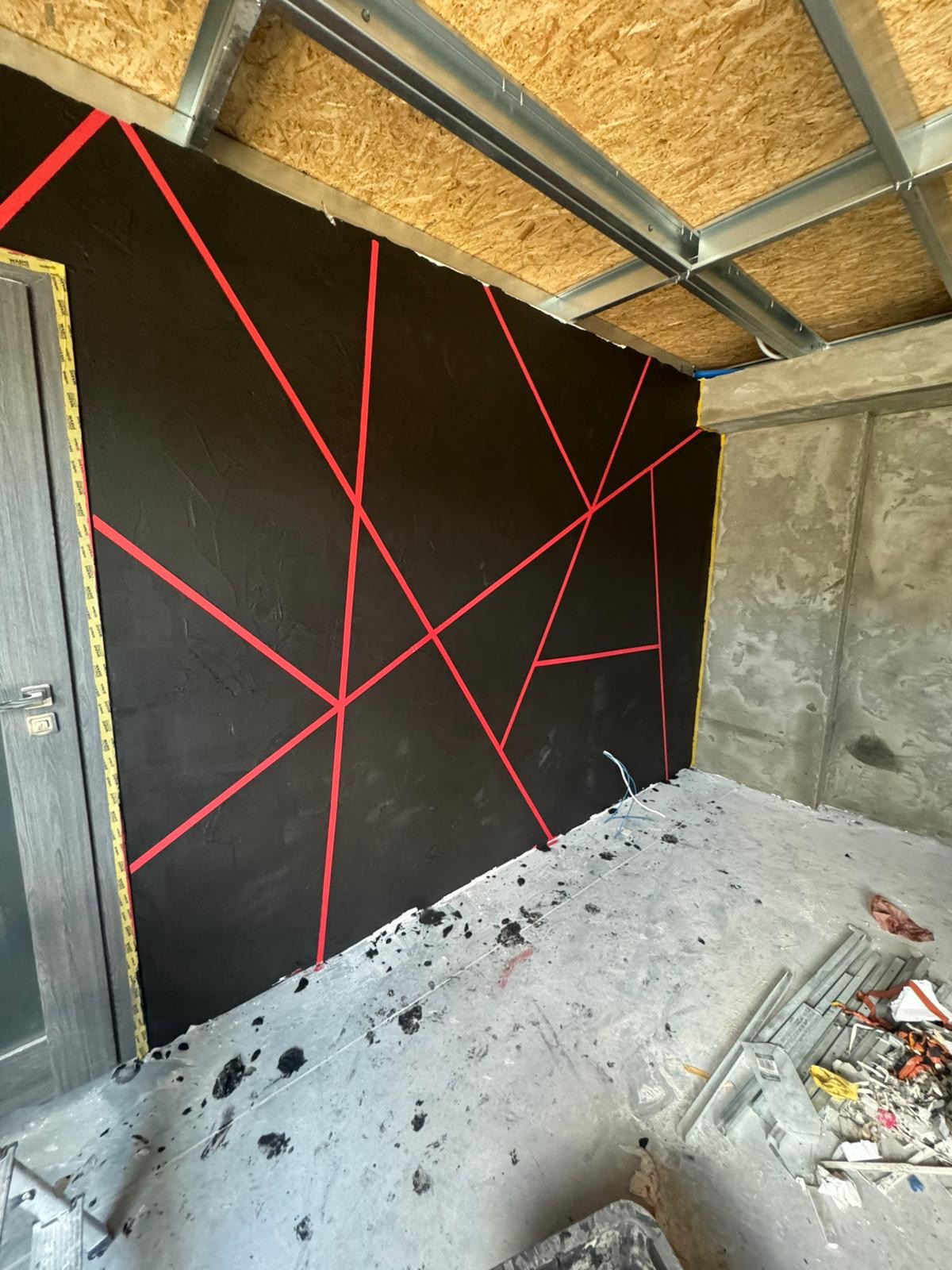 Czarna ściana z czerwoną, geometryczną dekoracją z taśmy malarskiej, surowy sufit z metalową konstrukcją i fragmentem płyty OSB, betonowa podłoga w trakcie prac.