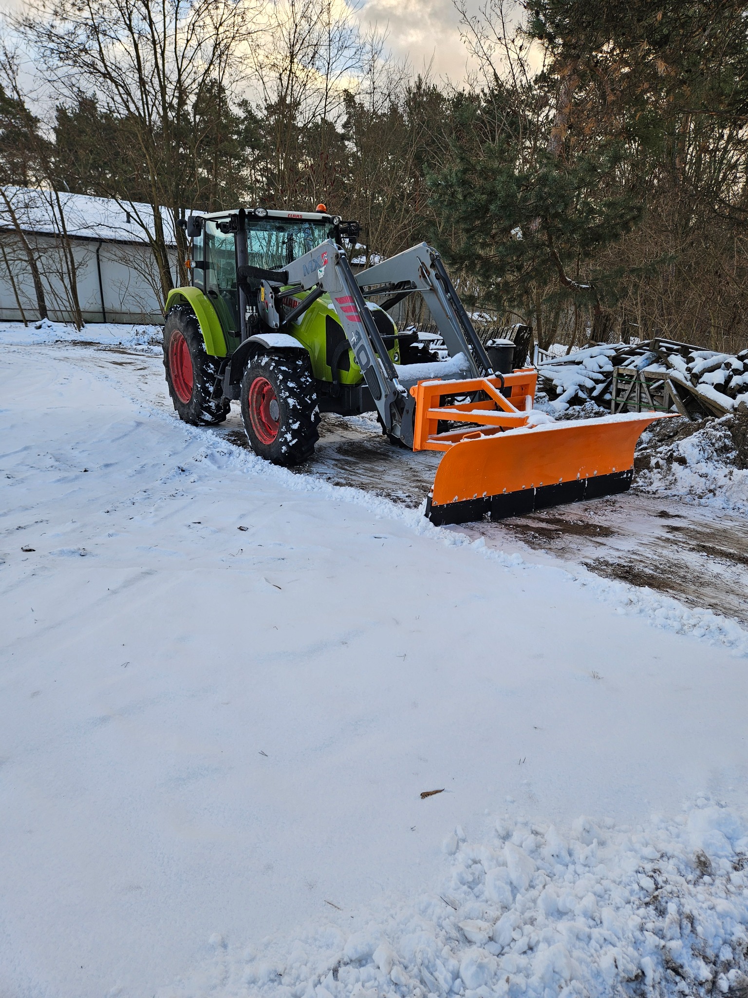 Zielony traktor Claas z pomarańczowym pługiem do śniegu na ośnieżonym terenie, w tle drzewa i budynek. Widok z perspektywy lekko z dołu.