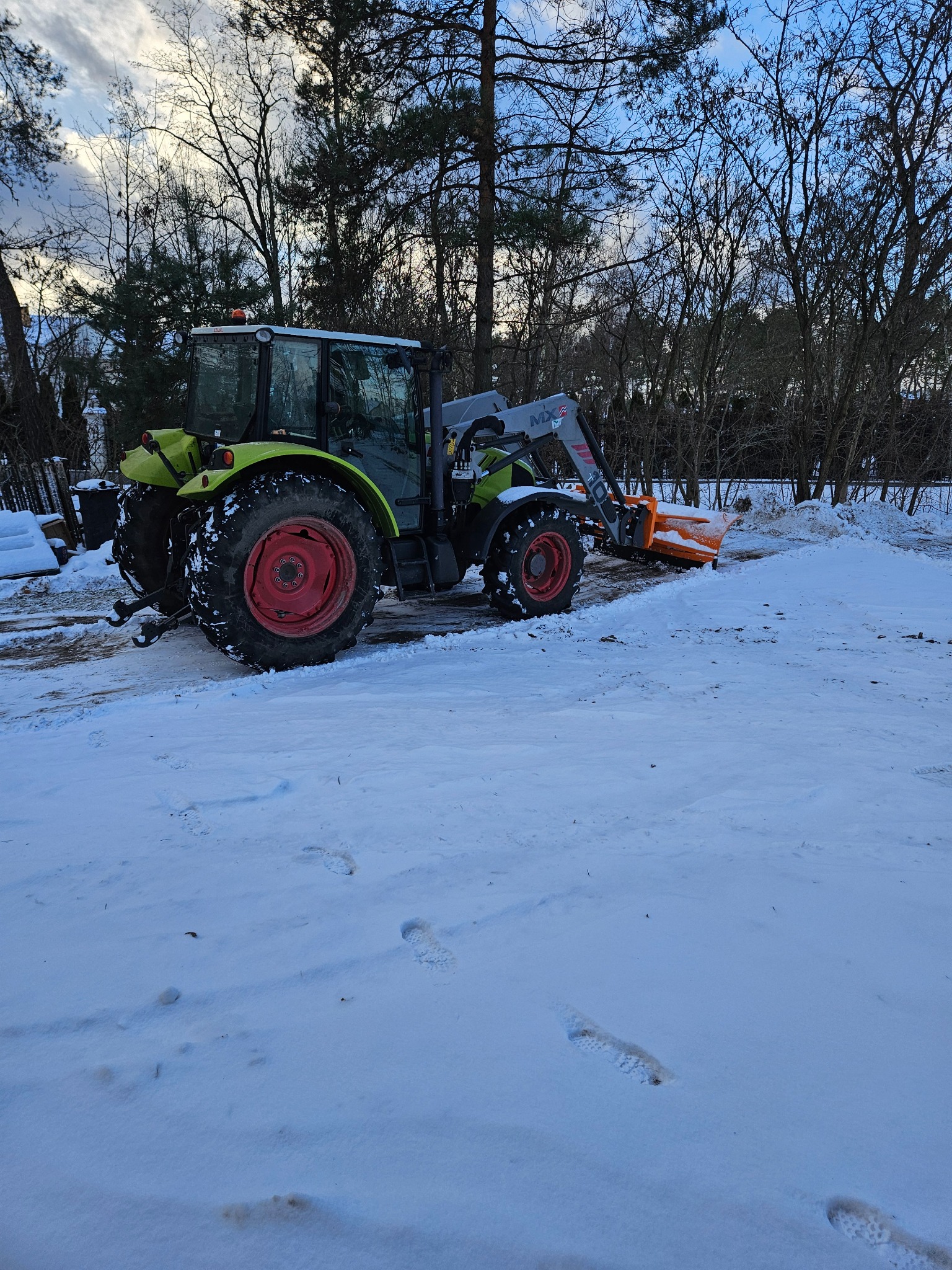 Zielony traktor Claas z pomarańczowym pługiem odśnieża zasypany śniegiem teren. Widoczne ślady opon i butów na śniegu, drzewa w tle.