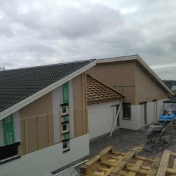 Cupryś Wojciech Bolisęga - Budowa domu z widoczną konstrukcją dachu z łatami, częściowo pokrytego czarną dachówką. Ściany w trakcie ocieplania, widoczna membrana i listwy. Szare niebo.