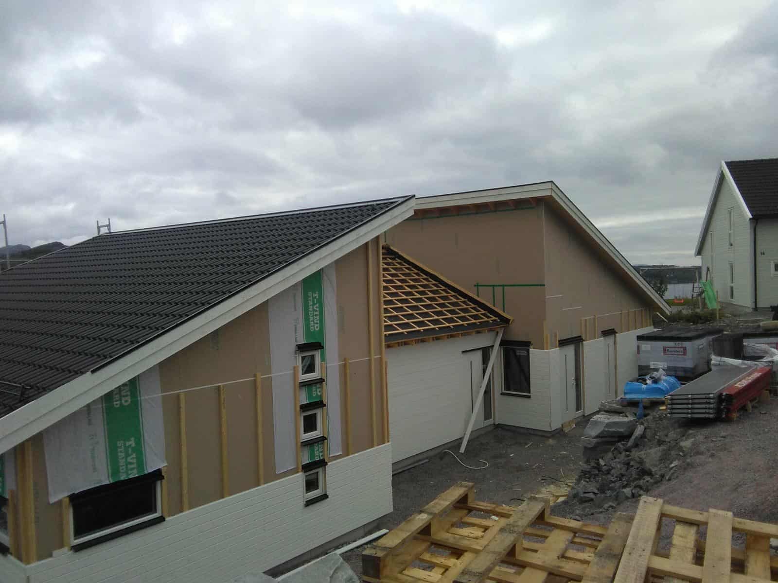 Budowa domu z widoczną konstrukcją dachu z łatami, częściowo pokrytego czarną dachówką. Ściany w trakcie ocieplania, widoczna membrana i listwy. Szare niebo.