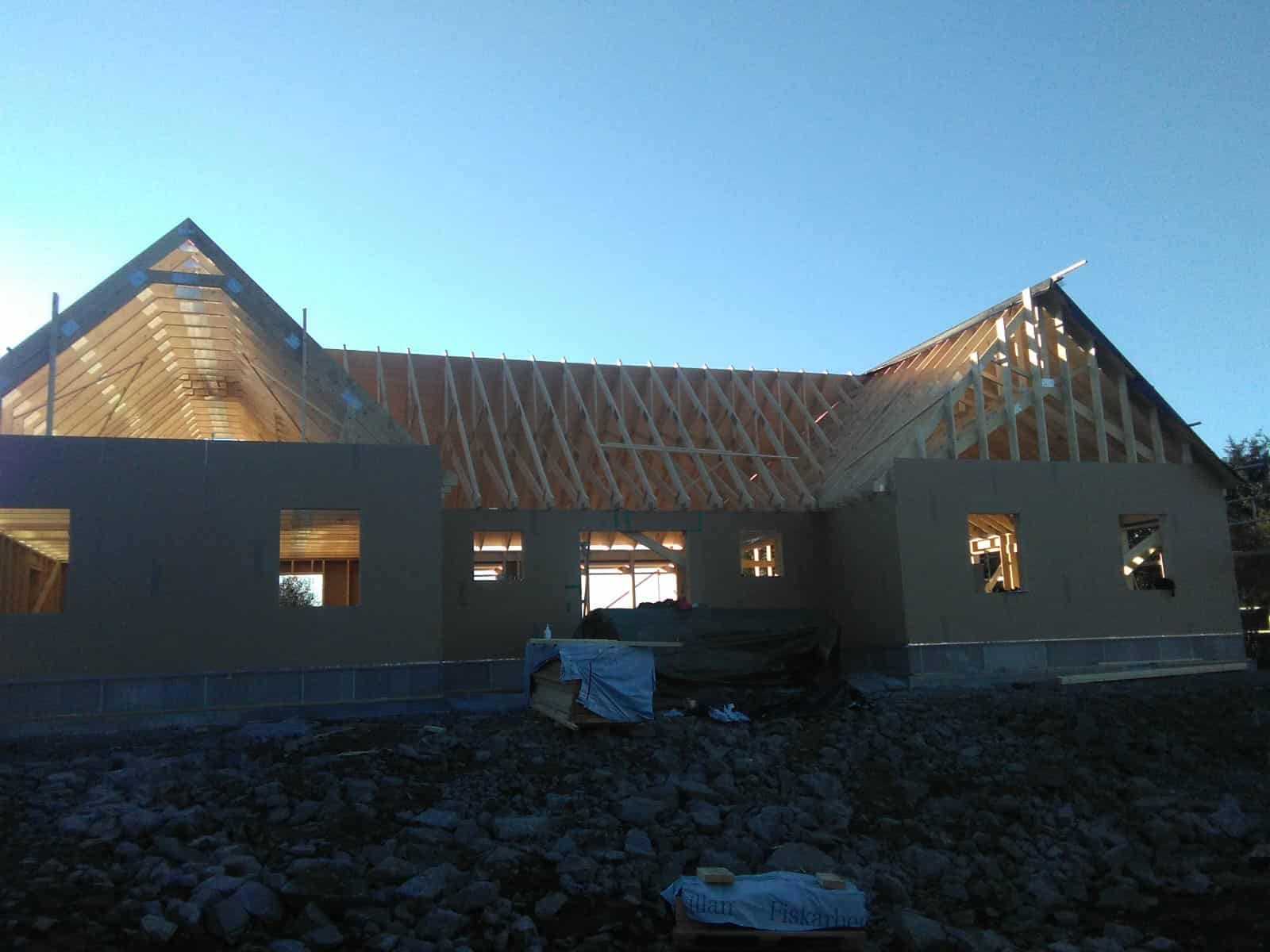 Budowa domu jednorodzinnego w stanie surowym otwartym: widoczna konstrukcja więźby dachowej z drewnianych belek, ściany szczytowe z otworami okiennymi, fundament.