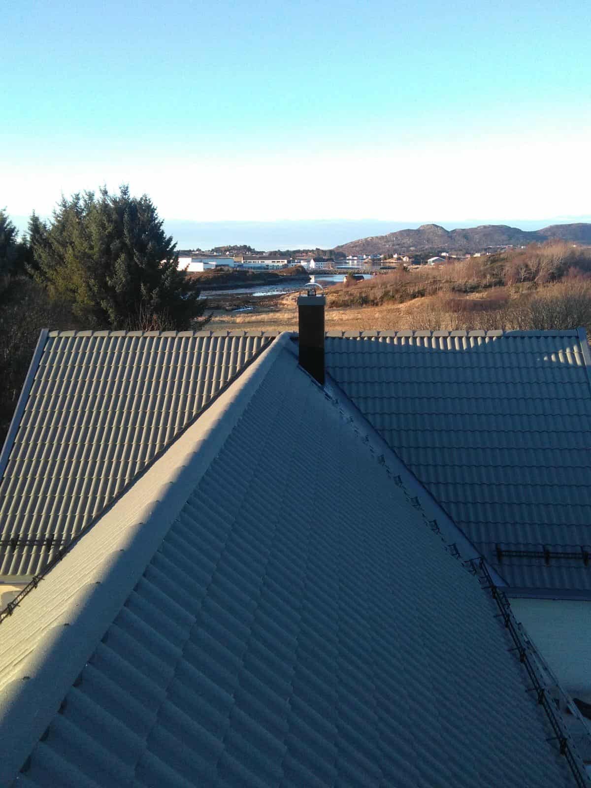 Szary dach dwuspadowy z kominem, widok z góry na tle krajobrazu z wodą i wzgórzami w oddali, w słoneczny dzień.