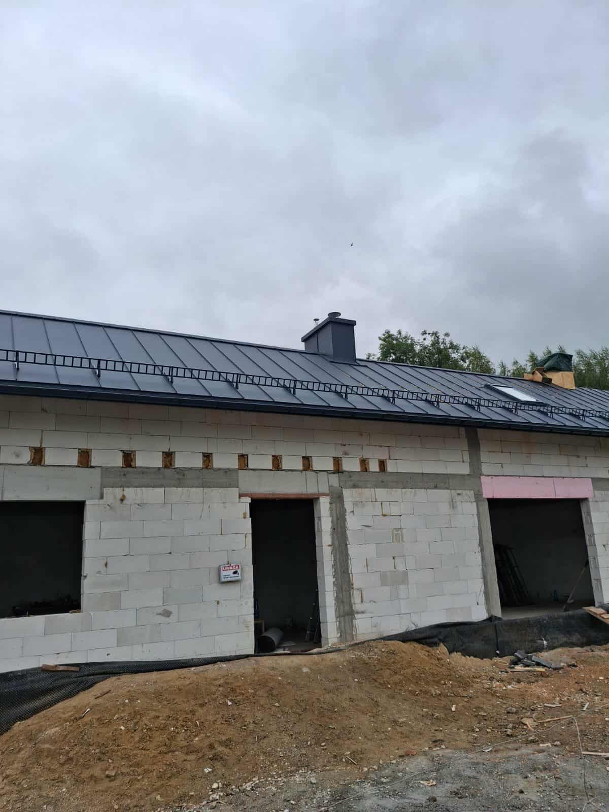 Szary dach z blachy na budynku w trakcie budowy, widoczne okna dachowe i komin. Ściany z białych bloczków, częściowo ocieplone. Teren wokół budynku przygotowany.