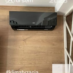 ARSEM sp z o.o. klimatyzacja, chłodnictwo, pompy ciepła, rekuperacja, wentylacja - Czarny klimatyzator AUX zamontowany na ścianie z drewnianą okładziną. Widoczny fragment białej konstrukcji i logo firmy Arsem. Na podłodze hashtag #klimatyzacja.