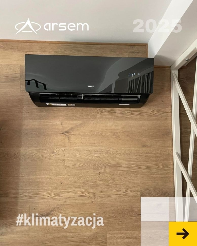 Czarny klimatyzator AUX zamontowany na ścianie z drewnianą okładziną. Widoczny fragment białej konstrukcji i logo firmy Arsem. Na podłodze hashtag #klimatyzacja.