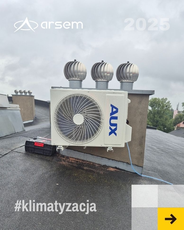 Biały klimatyzator AUX na dachu budynku z trzema kominami wentylacyjnymi. Obok skrzynka narzędziowa. Widoczny hashtag #klimatyzacja.