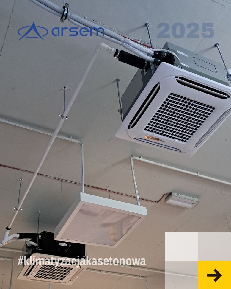 Kasetonowa klimatyzacja sufitowa z widocznymi rurami i instalacją, zamontowana w pomieszczeniu z oświetleniem. Ujęcie z dołu, widoczny fragment logo firmy.
