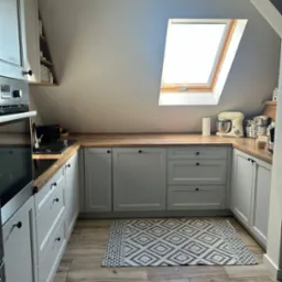 Jasna kuchnia na poddaszu w Twardogórze. Szare szafki z drewnianym blatem, okno dachowe i wzorzysty dywanik. Ustawna zabudowa kuchenna.