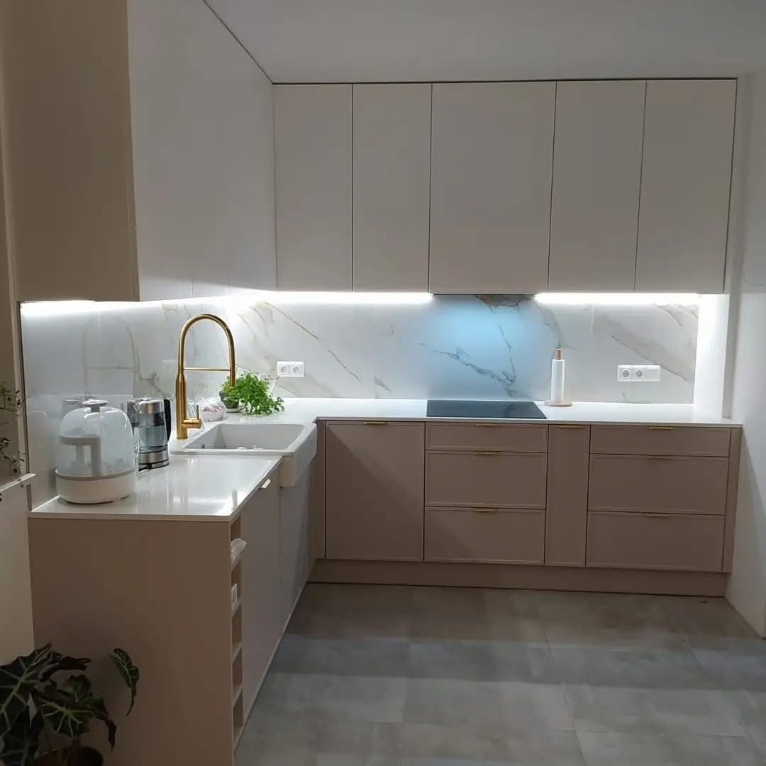 Jasna kuchnia z marmurowym backsplashiem i złotą armaturą. Górne szafki białe, dolne w odcieniach beżu. Oświetlenie LED pod szafkami. Minimalistyczny design.