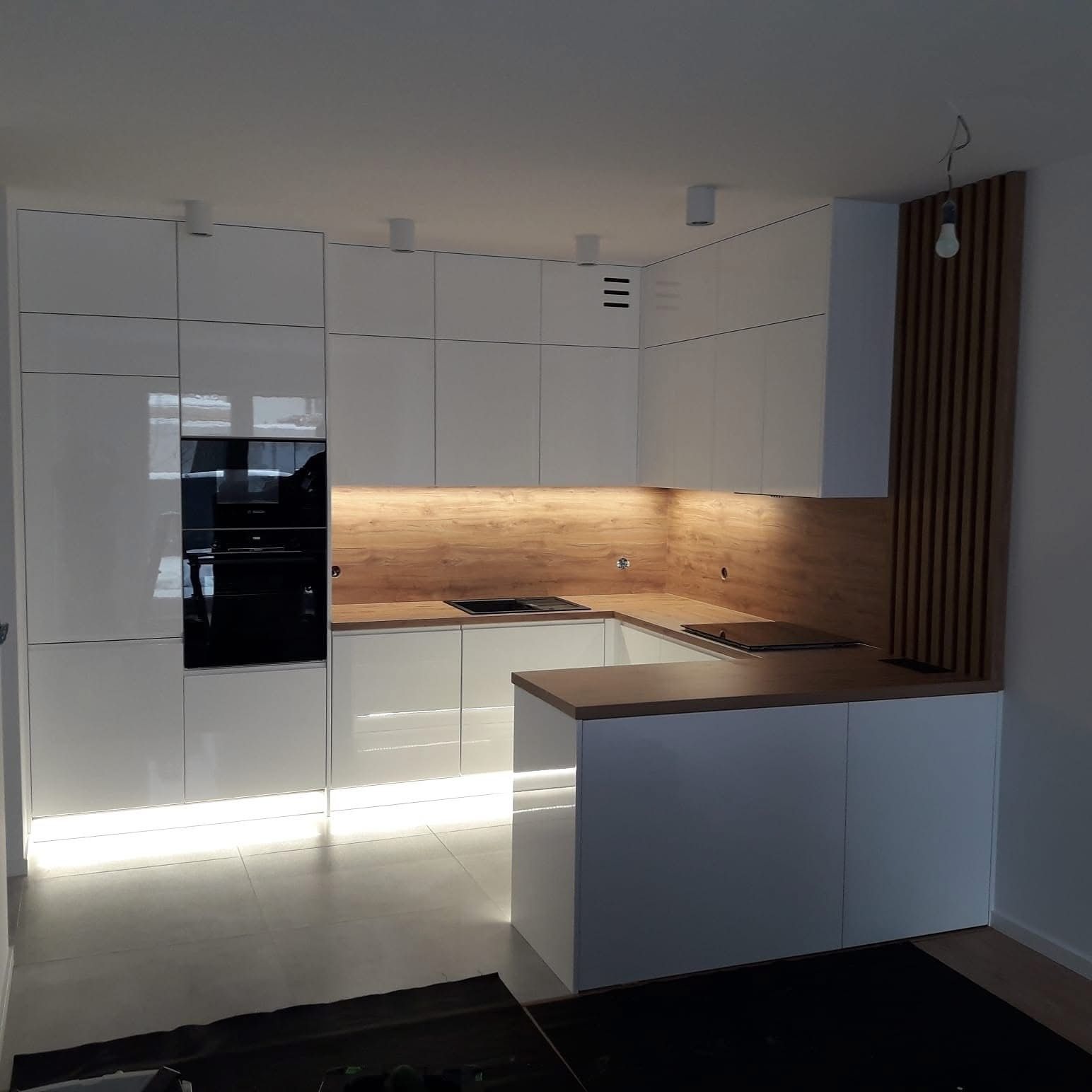 Nowoczesna kuchnia z białymi, lakierowanymi frontami, drewnianym blatem i oświetleniem LED pod szafkami. Na ścianie dekoracyjny panel z drewnianych listewek.