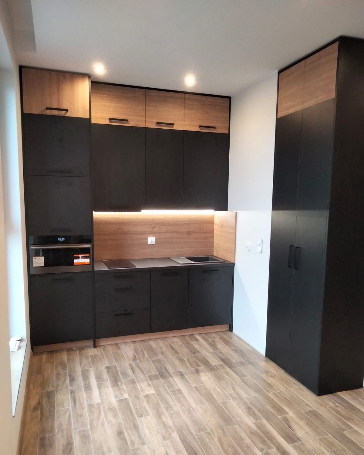 Nowoczesna kuchnia na wymiar: czarne matowe fronty, drewniane akcenty na górnych szafkach i panelu ściennym, oświetlenie LED podszafkowe, drewnopodobna podłoga.