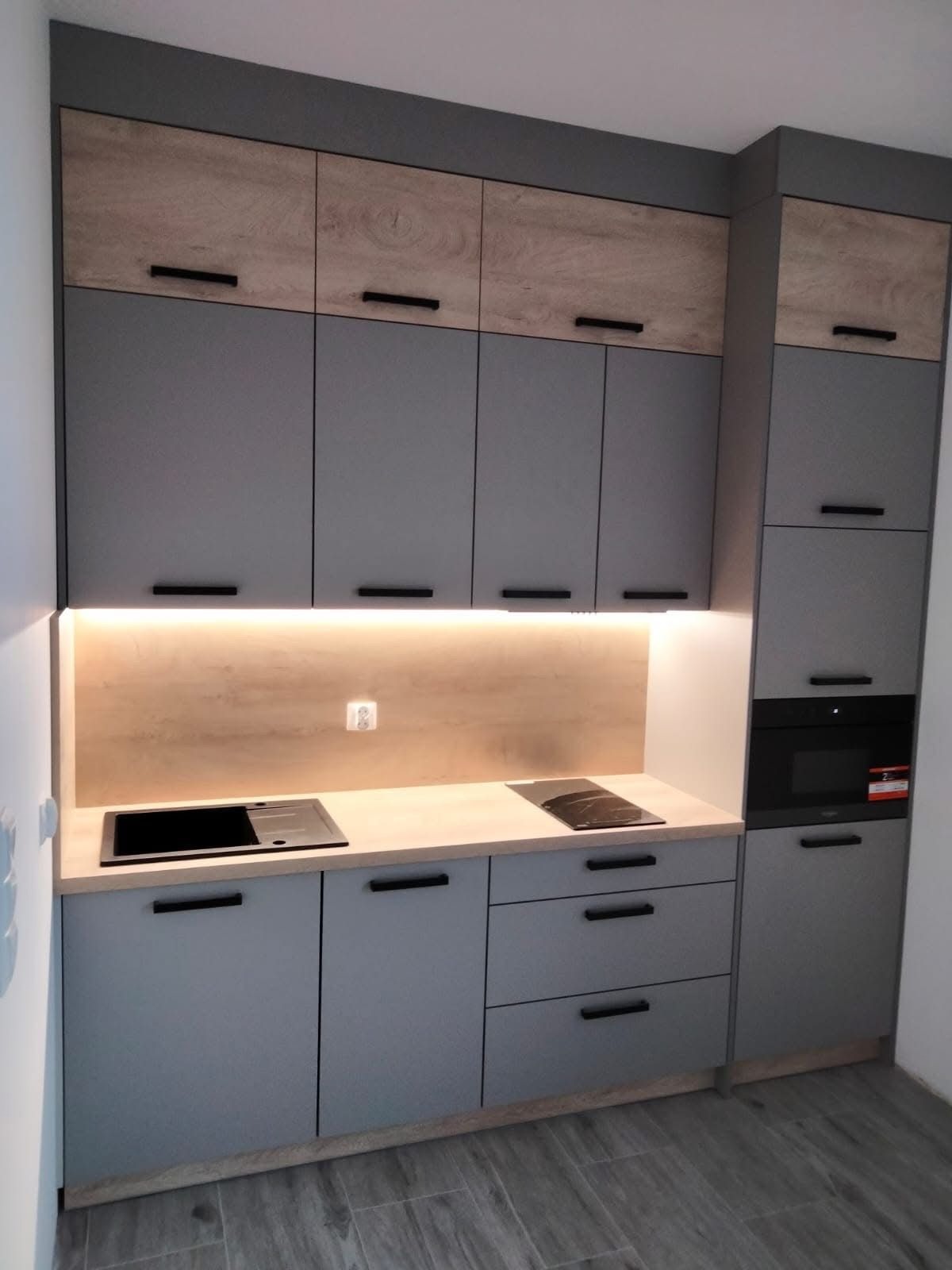 Szara zabudowa kuchenna z drewnianymi akcentami, czarnymi uchwytami i oświetleniem LED nad blatem. Widoczny zlew, płyta indukcyjna i piekarnik.