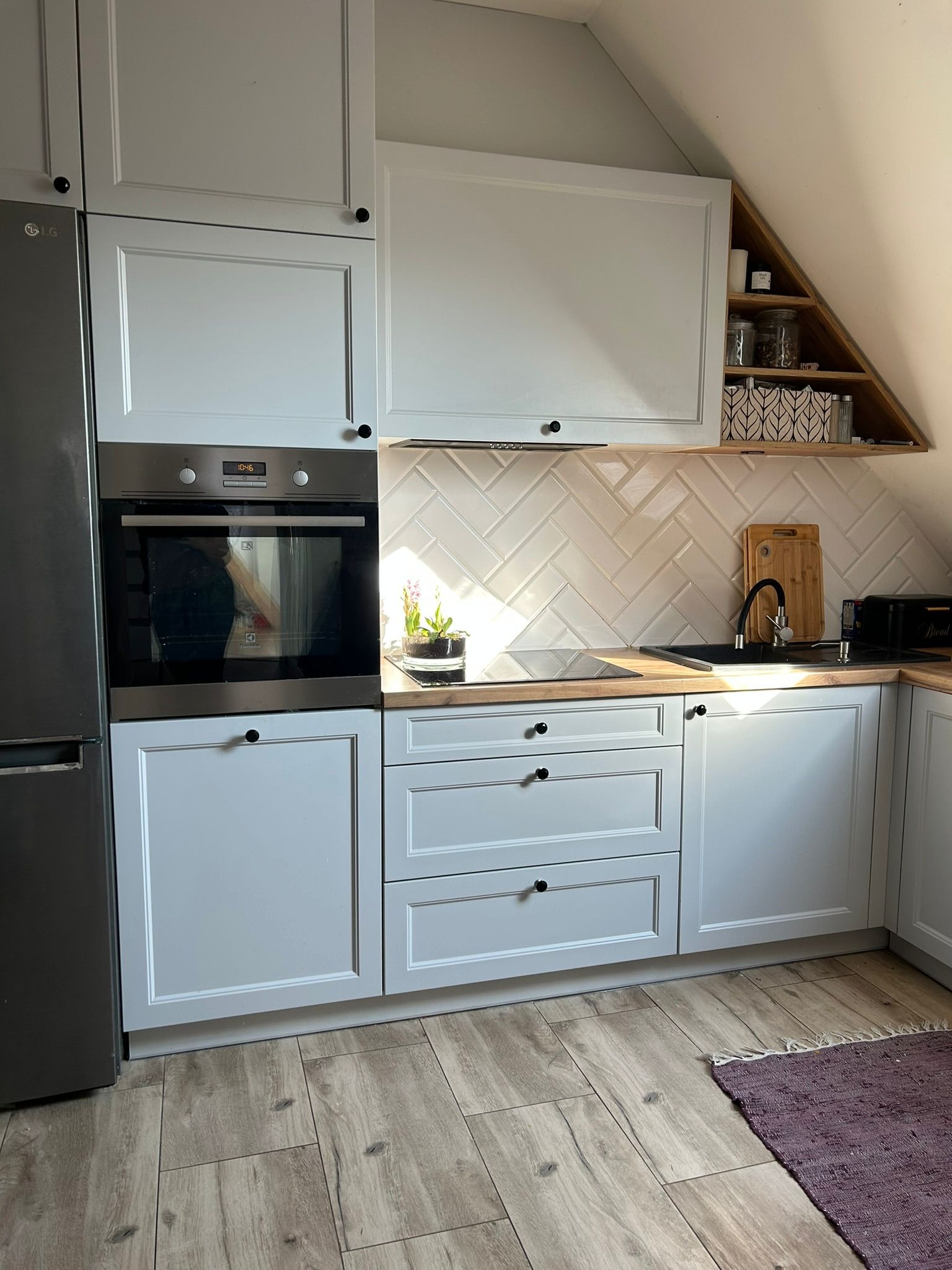 Jasna kuchnia na poddaszu z białymi szafkami, czarnymi uchwytami i drewnianym blatem. Uwagę przykuwają płytki ułożone w jodełkę i trójkątna półka.