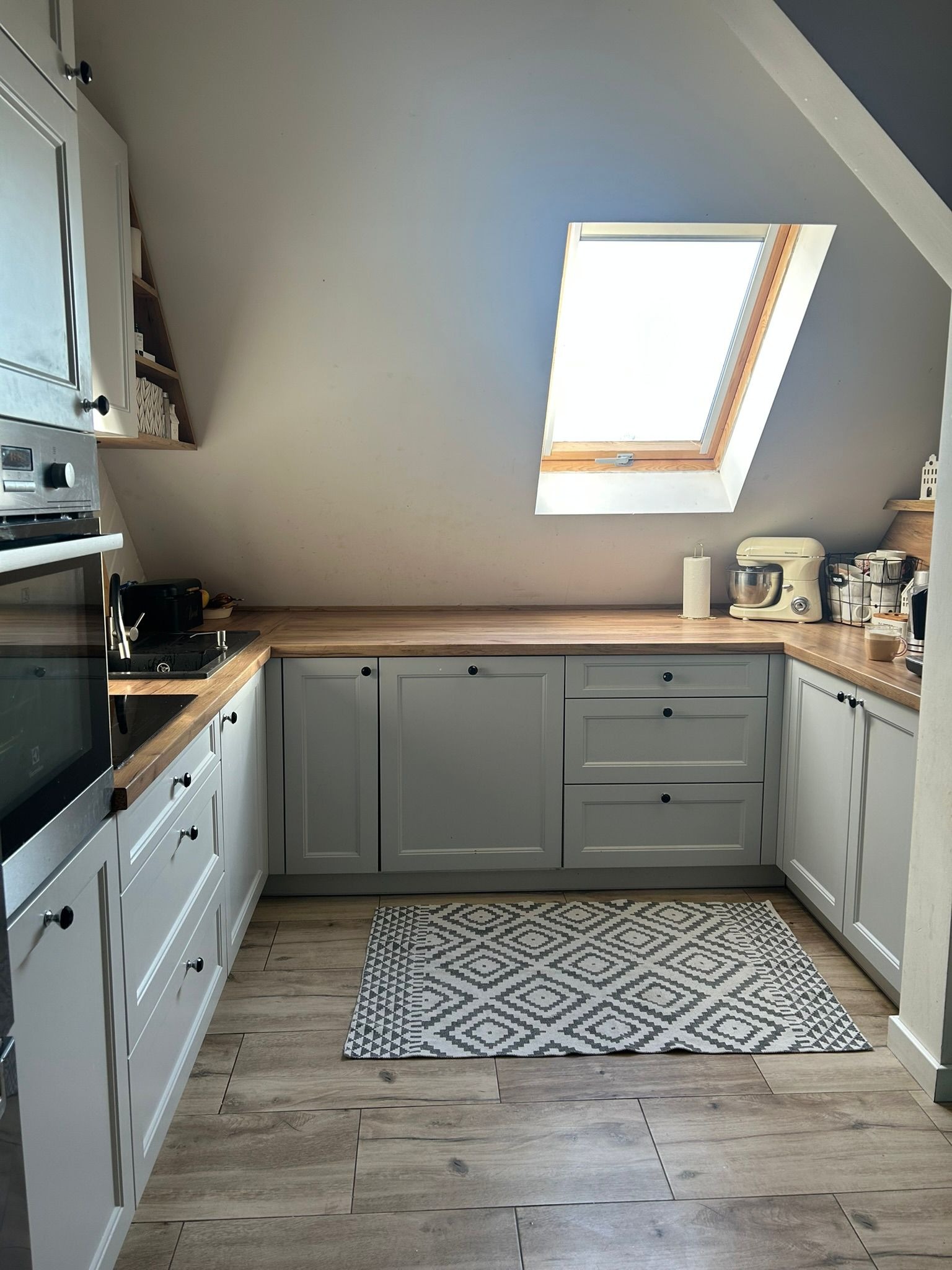 Jasna kuchnia na poddaszu w Twardogórze. Szare szafki z drewnianym blatem, okno dachowe i wzorzysty dywanik. Ustawna zabudowa kuchenna.