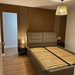 MV EliteX Mykhailo Volovetskyi - Pokój z beżowym łóżkiem i panelami ściennymi w odcieniach brązu. Widoczna lampa wisząca z abażurem z naturalnego materiału i drewniana podłoga.