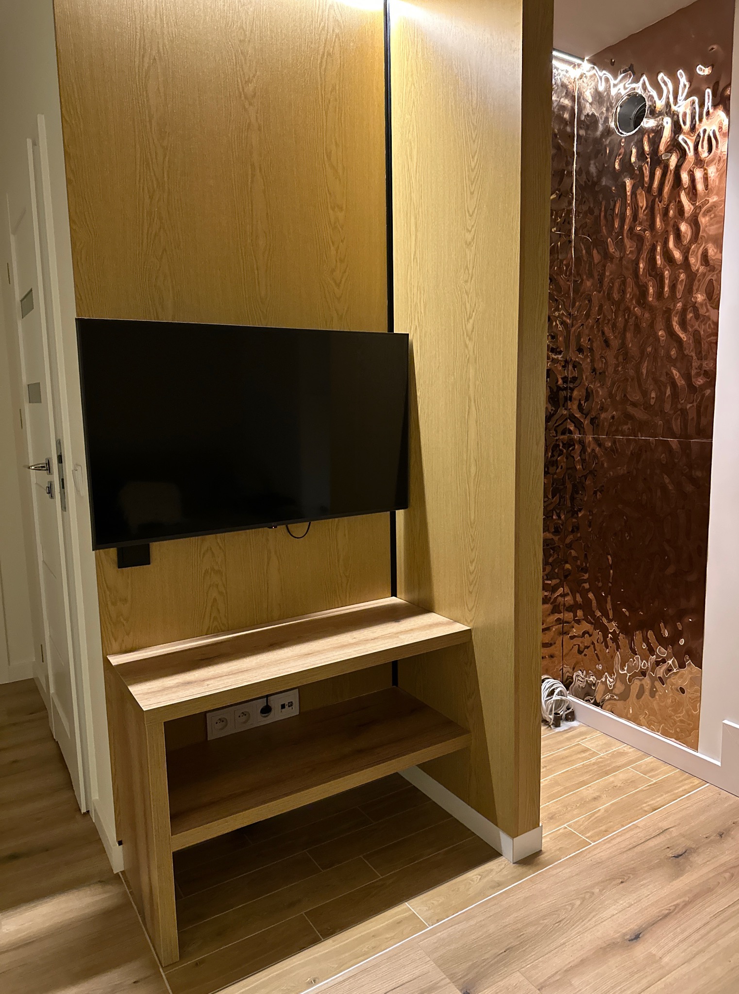 Nowoczesny salon z drewnianą zabudową TV, półką i dekoracyjną ścianą z miedzianych paneli. Widoczne gniazdka elektryczne i fragment podłogi z płytek imitujących drewno.