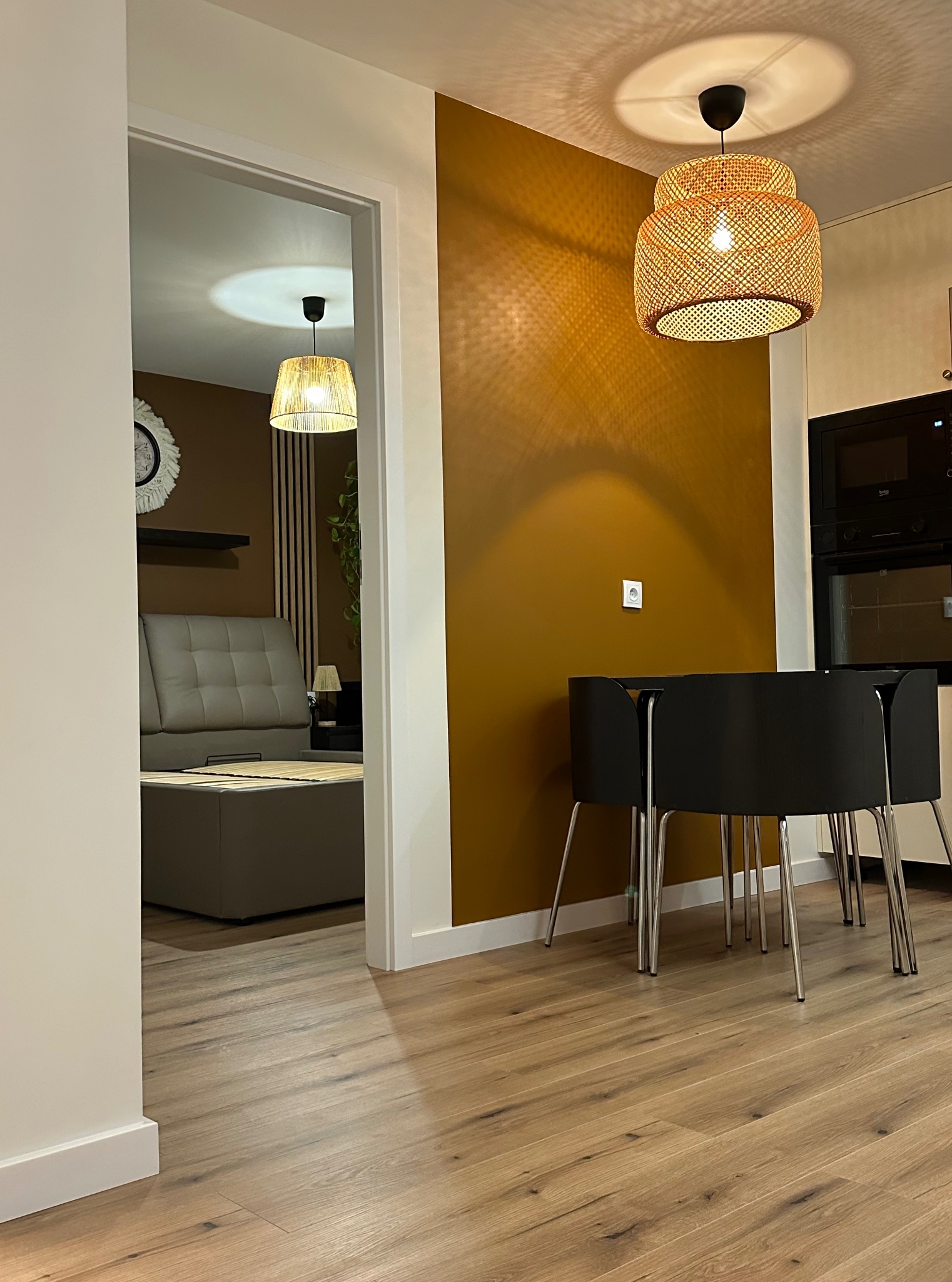 Nowoczesne wnętrze: parkiet, musztardowa ściana, czarne krzesła i lampa z rattanu. Widok na salon z szarą sofą przez białe drzwi. Minimalistyczny design.