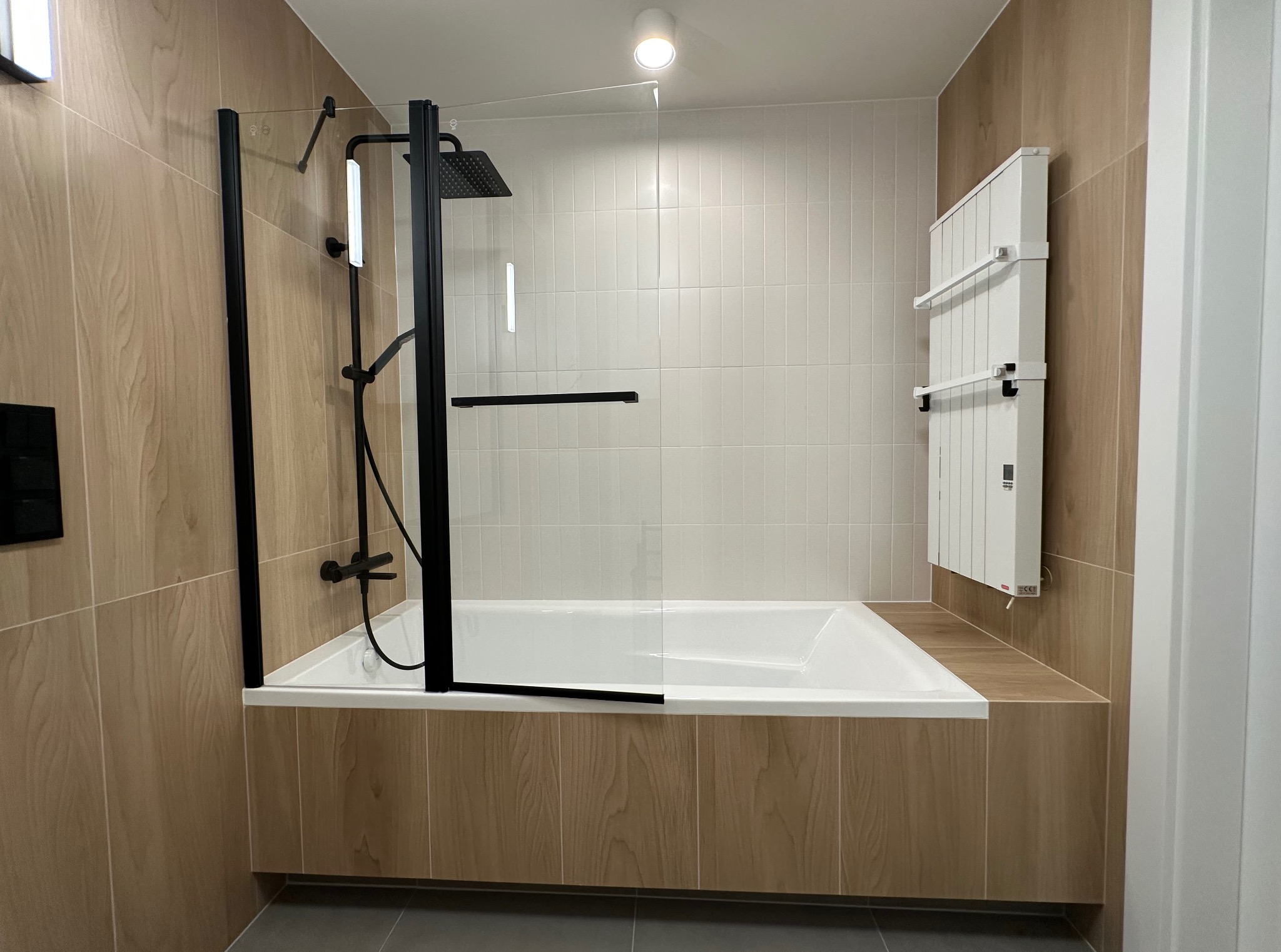 Nowoczesna łazienka po remoncie: biała wanna z czarną armaturą i szklanym parawanem, grzejnik drabinkowy, beżowe płytki imitujące drewno, szara podłoga.