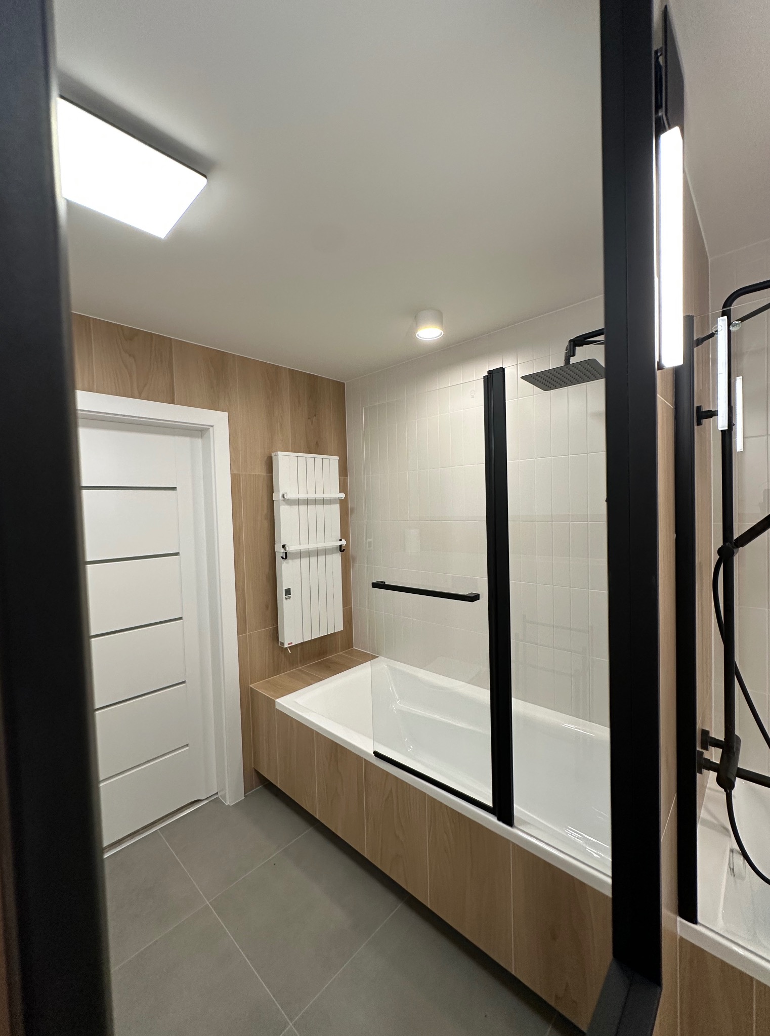 Nowoczesna łazienka po remoncie: biała wanna z czarną kabiną, drewnopodobne płytki, grzejnik drabinkowy i szara podłoga. Minimalistyczny design.