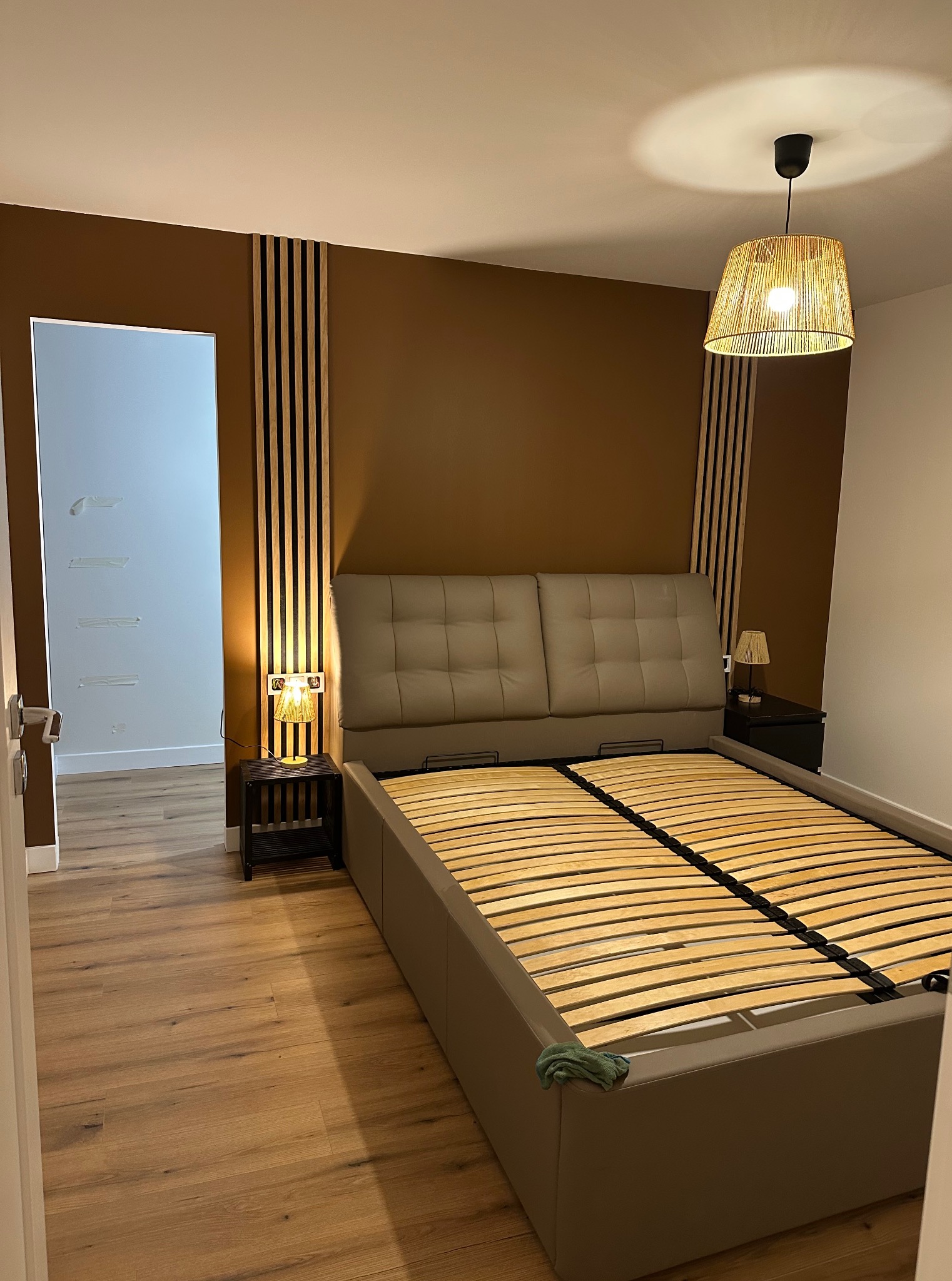 Pokój z beżowym łóżkiem i panelami ściennymi w odcieniach brązu. Widoczna lampa wisząca z abażurem z naturalnego materiału i drewniana podłoga.