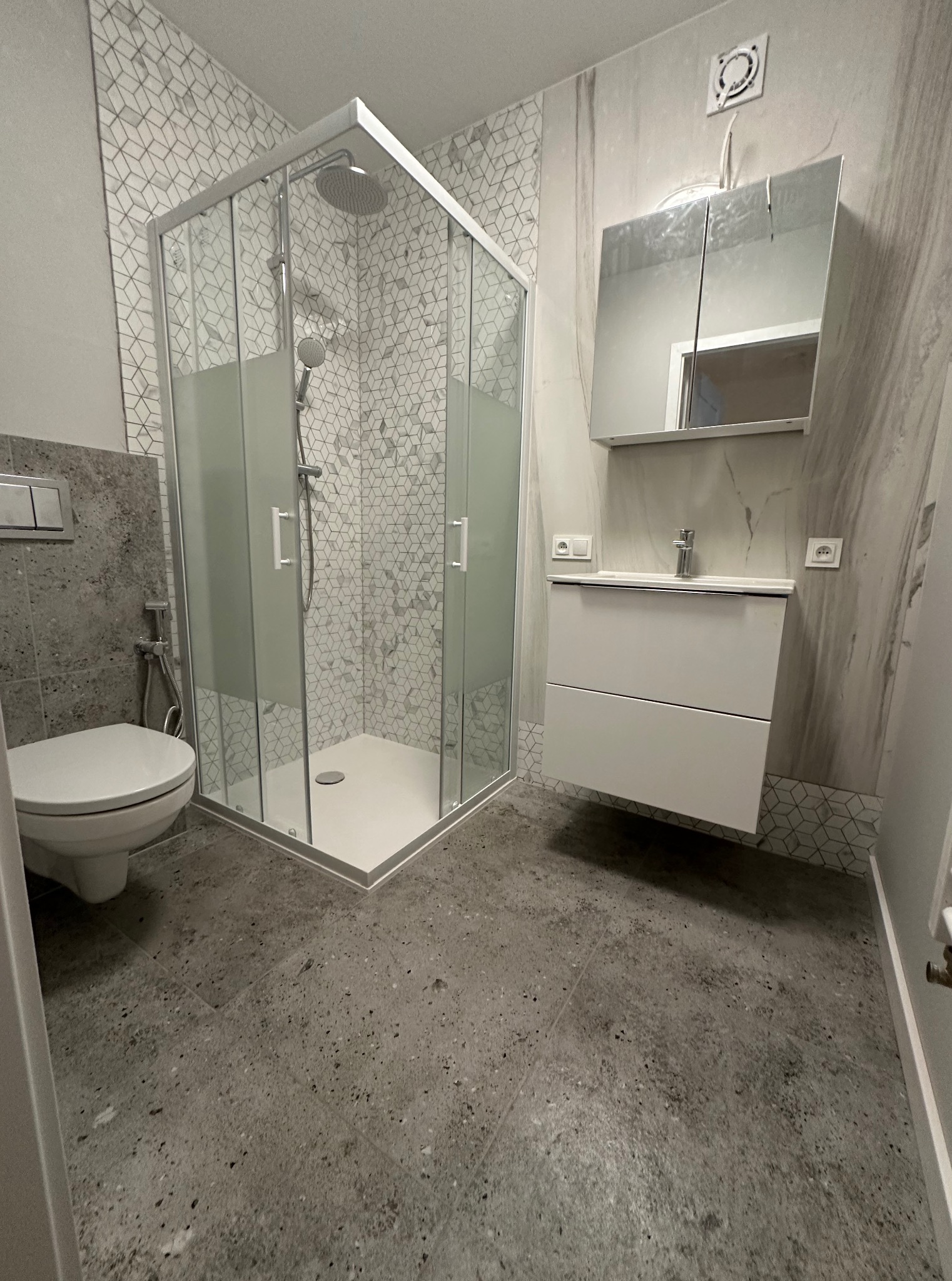 Nowoczesna łazienka po remoncie: kabina prysznicowa z geometrycznym wzorem płytek, umywalka z szafką i lustrem. Szare płytki podłogowe i częściowo na ścianach.