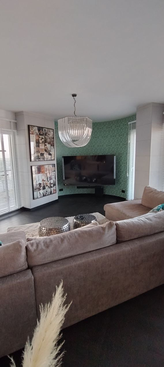 Salon z sofą w odcieniach beżu, srebrnymi stolikami i telewizorem na ścianie z zieloną tapetą. Nowoczesny żyrandol i dekoracje ścian dopełniają wystrój.