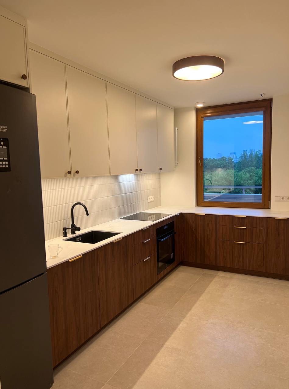 Nowoczesna kuchnia z drewnianymi szafkami dolnymi i białymi górnymi, czarnym zlewem i płytą indukcyjną. Duże okno z widokiem na zieleń. Minimalistyczne oświetlenie.